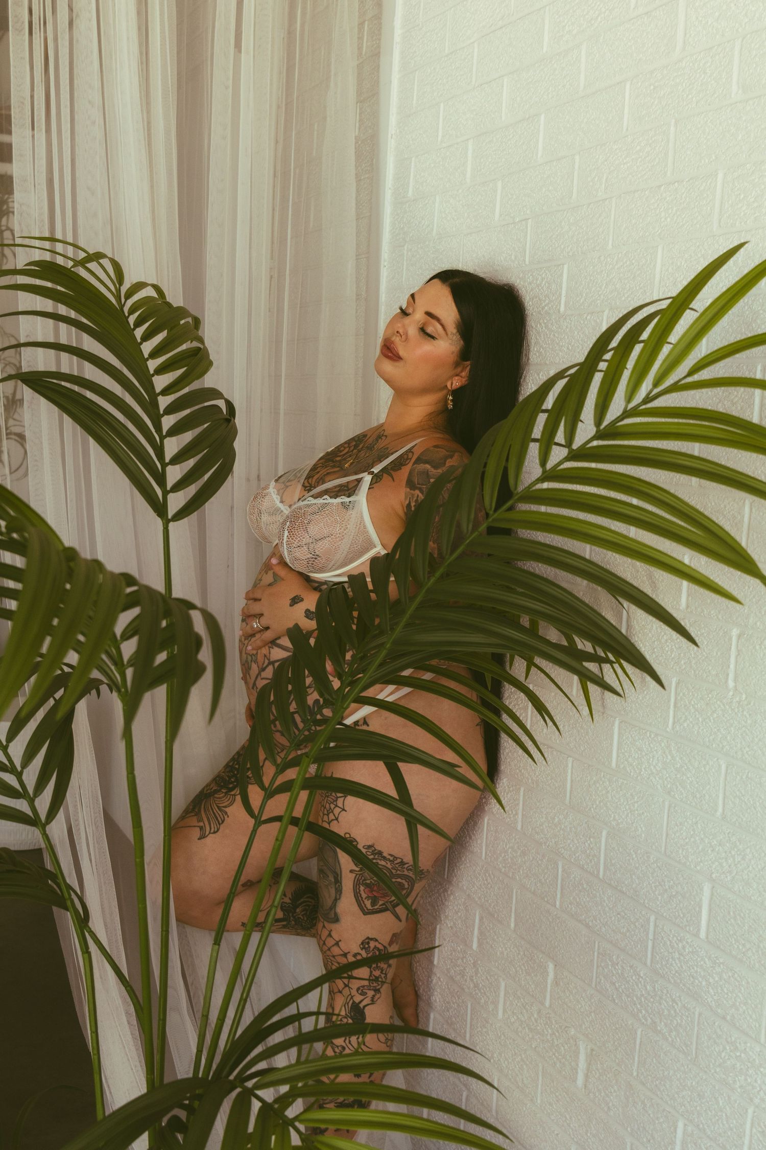 Tattooed pregnant woman in white lace lingerie posing beside tropical plants against a white brick wall.