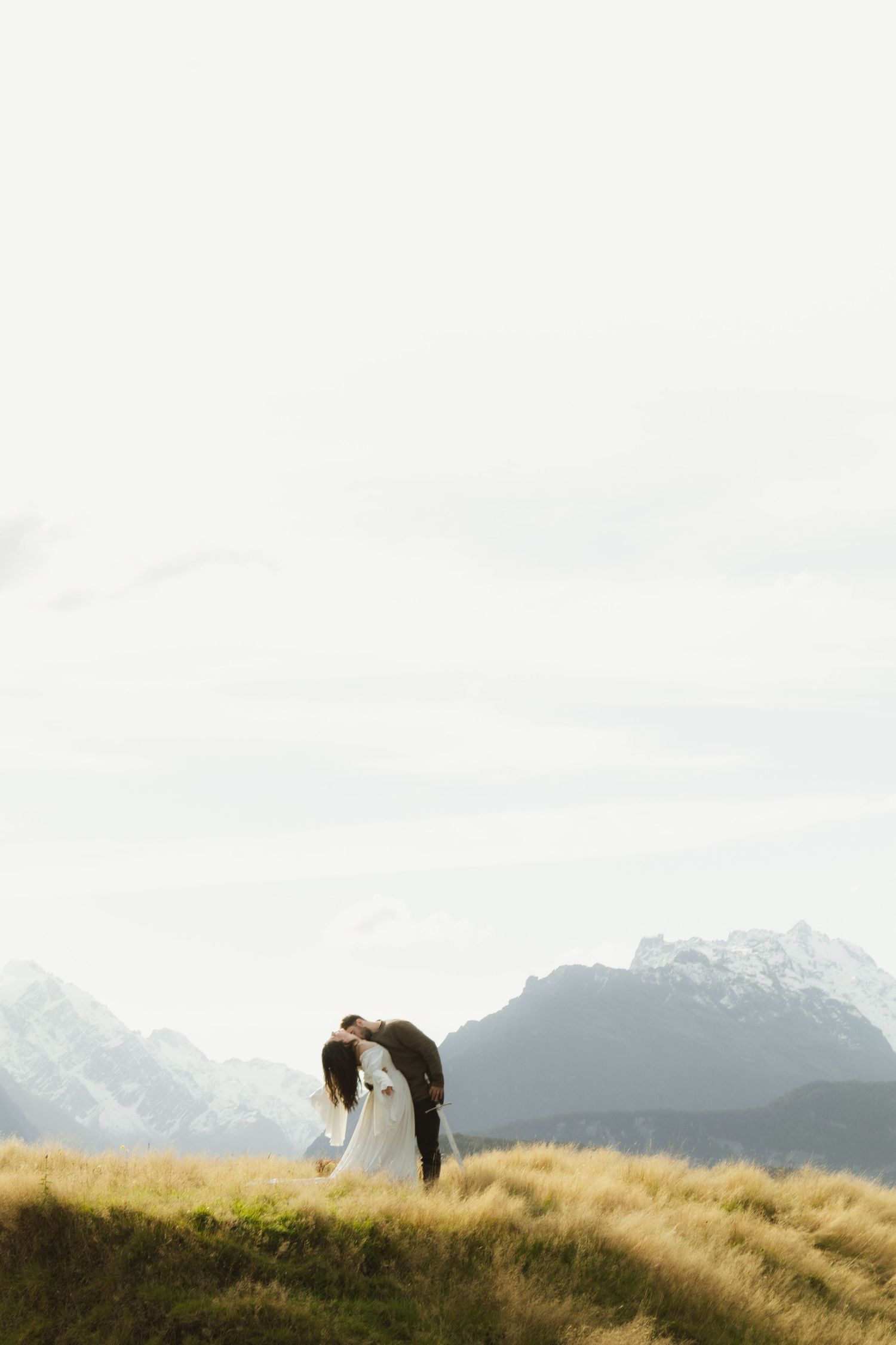 Bride and groom share a romantic kiss on a misty mountain meadow with snow-capped peaks in the background.