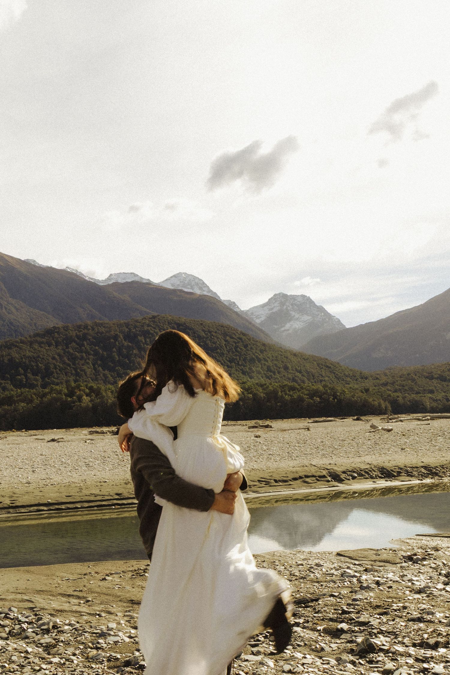Couple embracing in white wedding attire with stunning mountain landscape and reflective river in background.