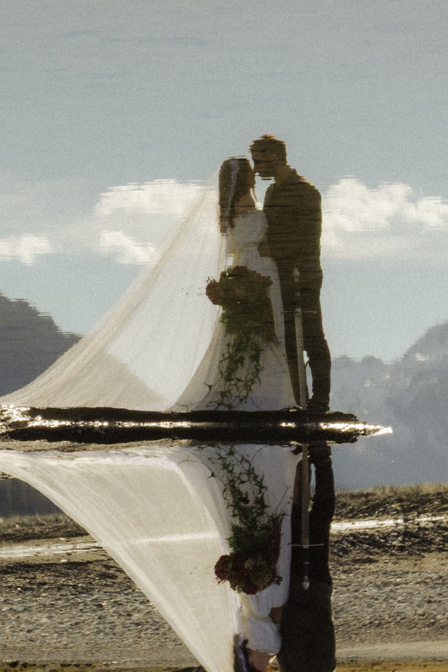 Bride and groom kissing with long veil flowing, reflected in still water with mountain backdrop.