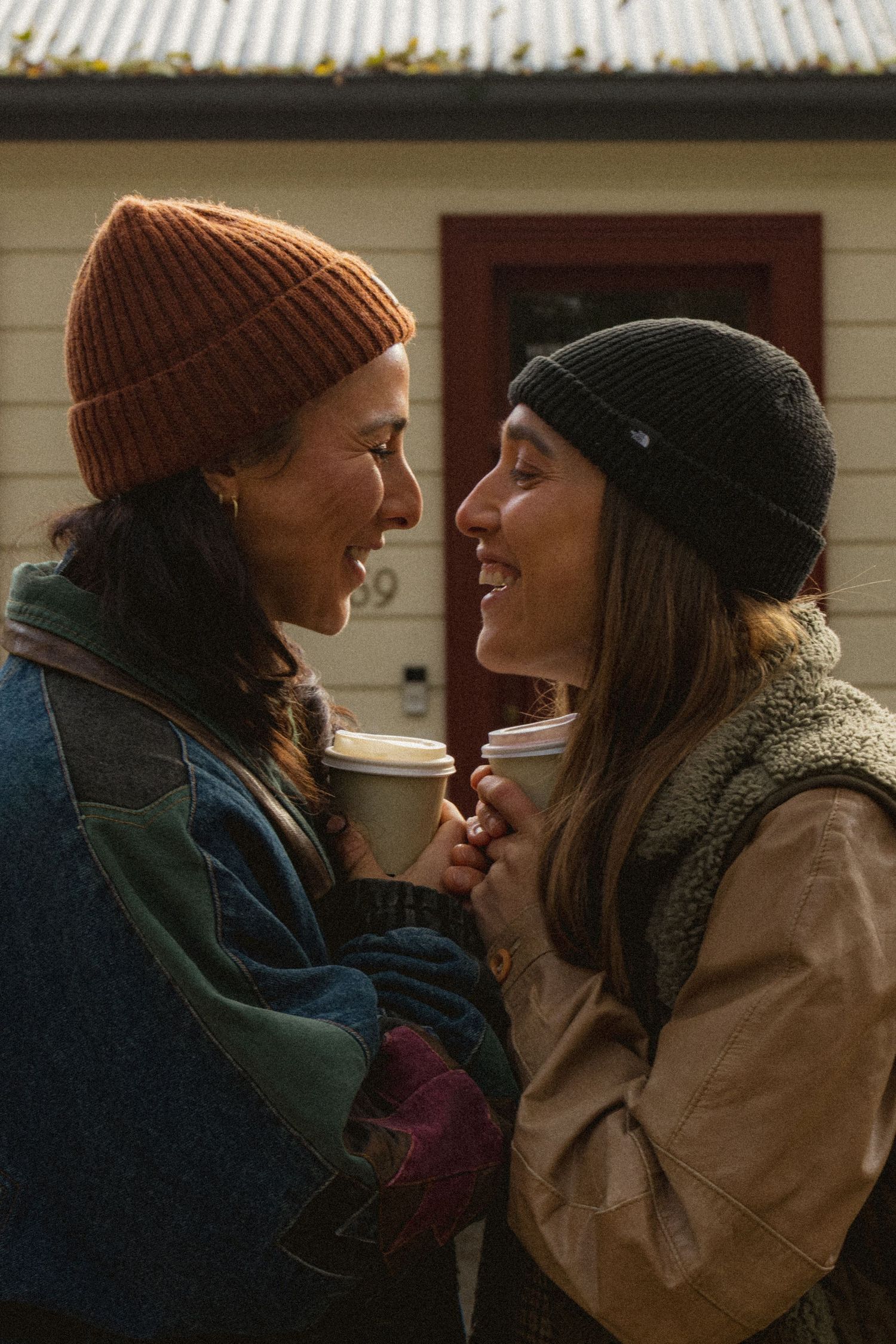 Two women in beanies share a joyful moment holding coffee cups outside a white house.
