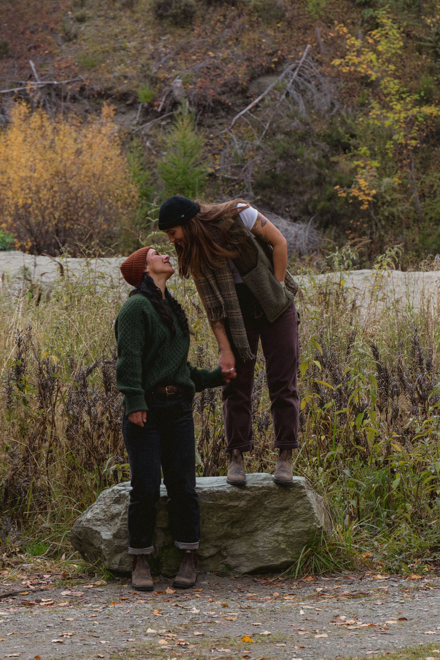 Couple sharing a romantic moment outdoors in fall, one standing on a rock holding hands with the other.