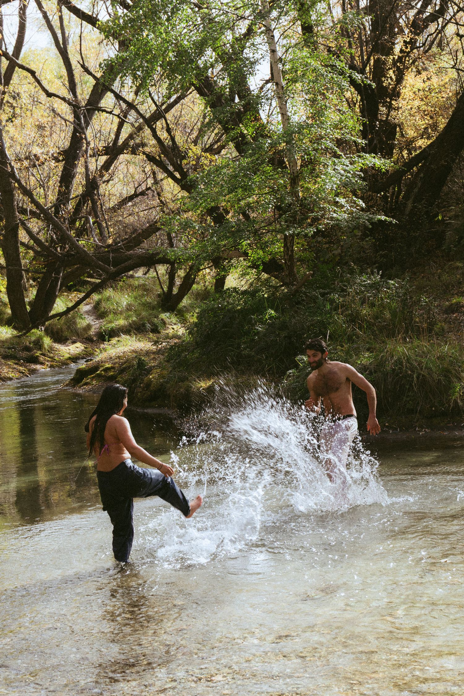 A couple splashing water in a shallow forest creek, surrounded by lush green trees on a sunny day.