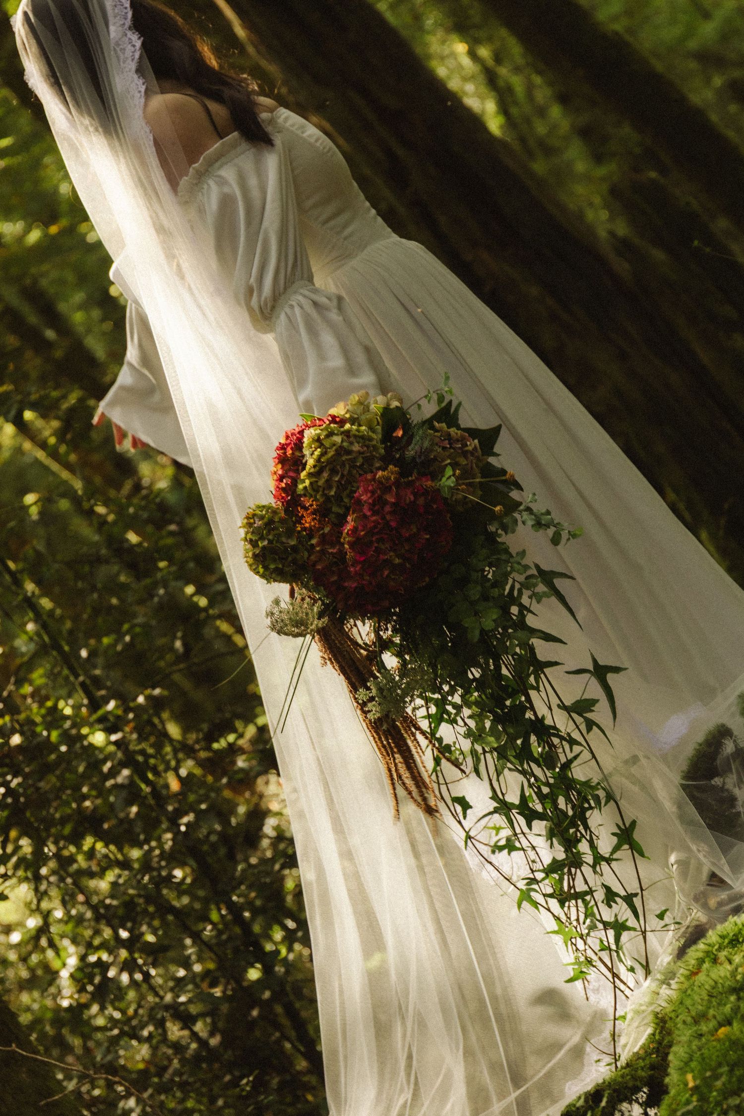 Bride in white gown and veil holds colorful bouquet with red hydrangeas and greenery in a sunlit forest setting.