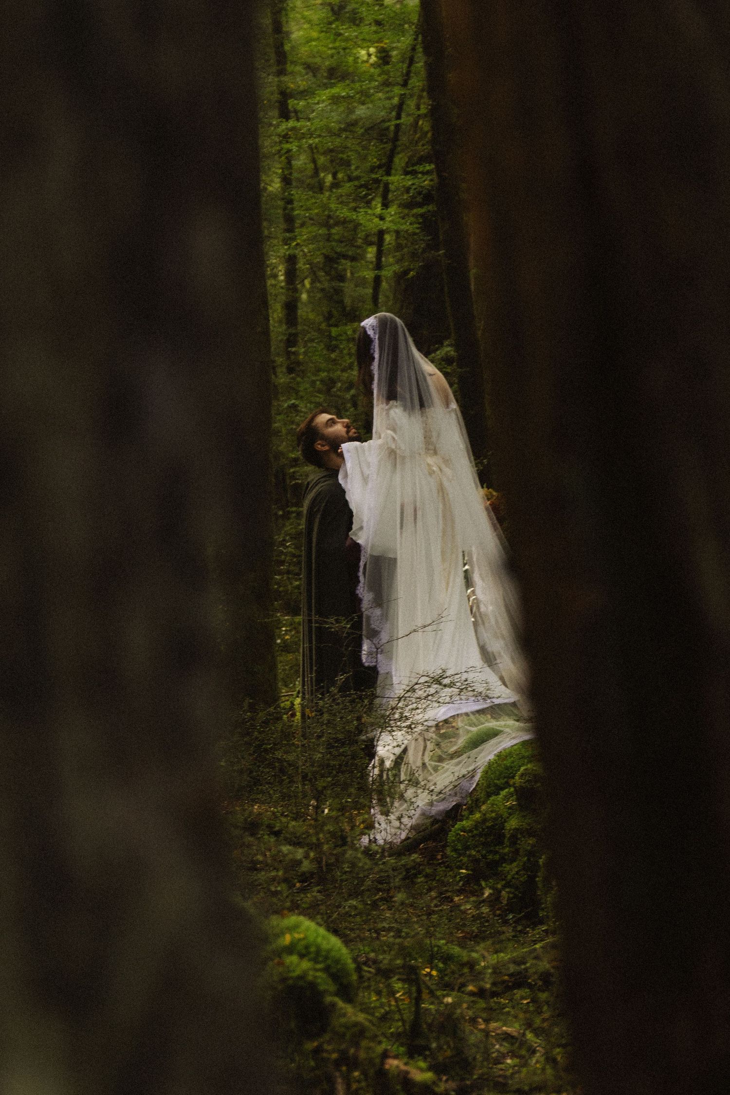 Bride and groom share romantic moment in lush green forest, bride wearing flowing white gown and veil.