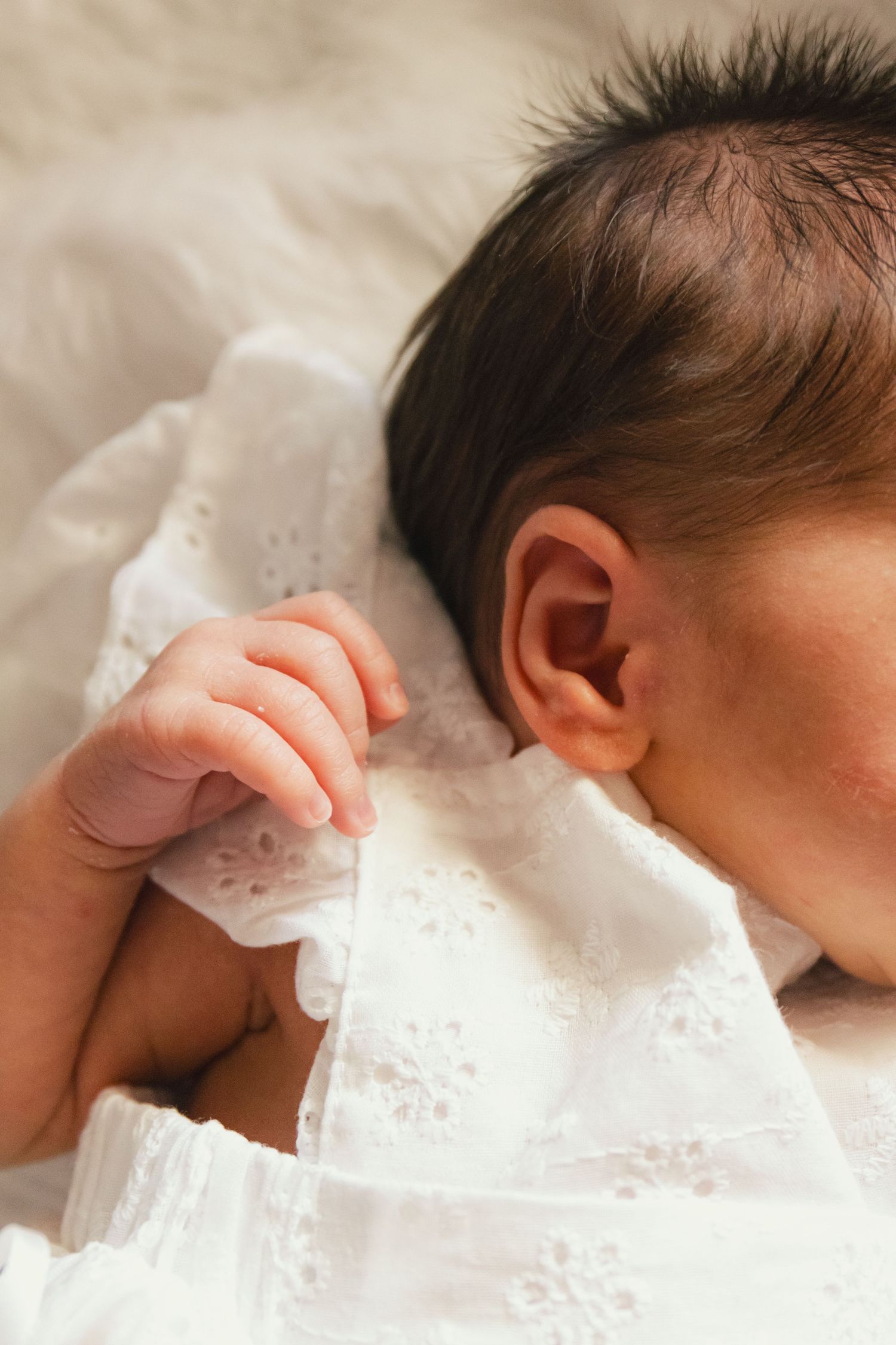 Close-up of a sleeping newborn baby with dark hair, wearing a white eyelet dress, tiny hand visible near ear.