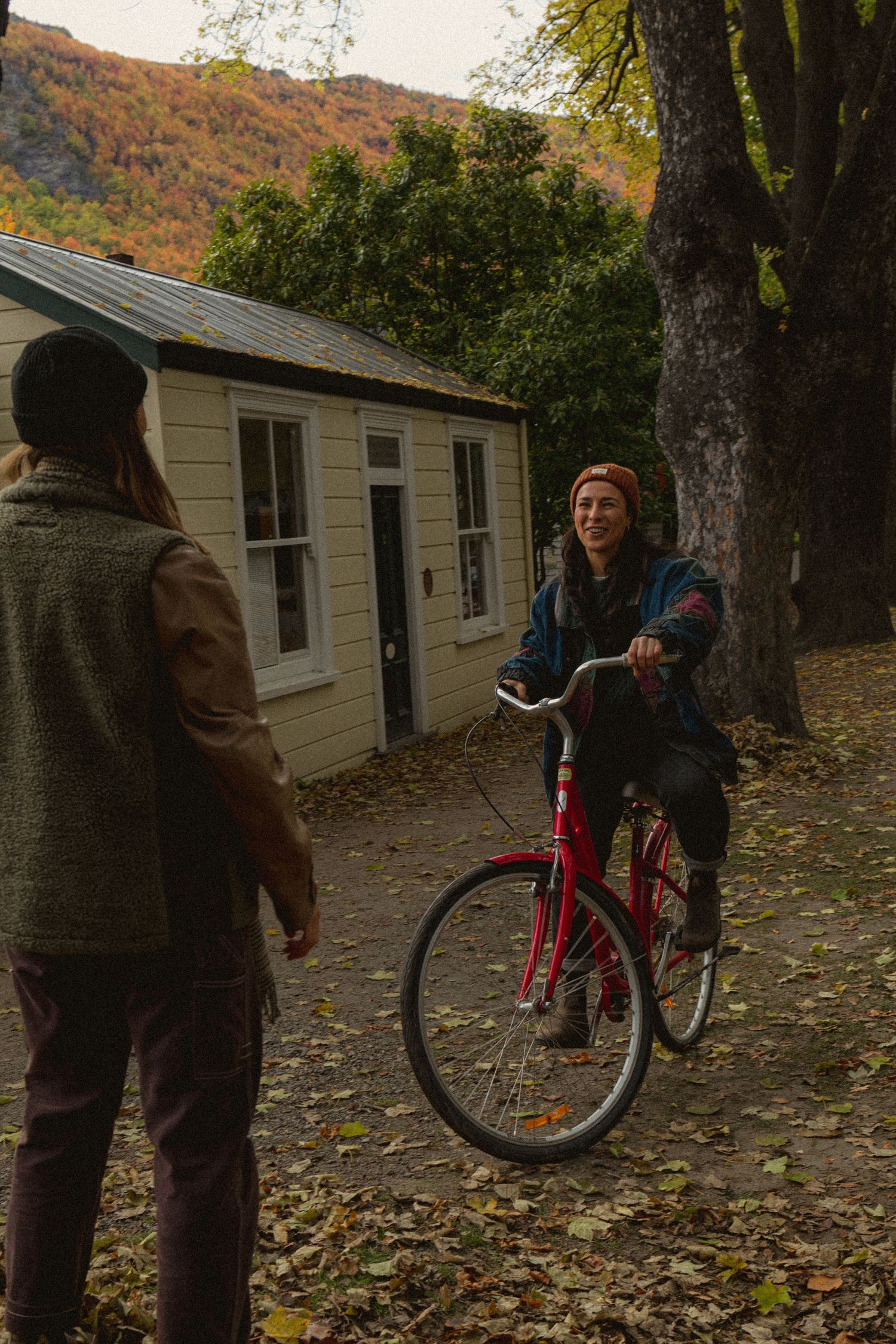 Person on red bicycle chatting with another near a cottage, surrounded by autumn trees and fallen leaves.