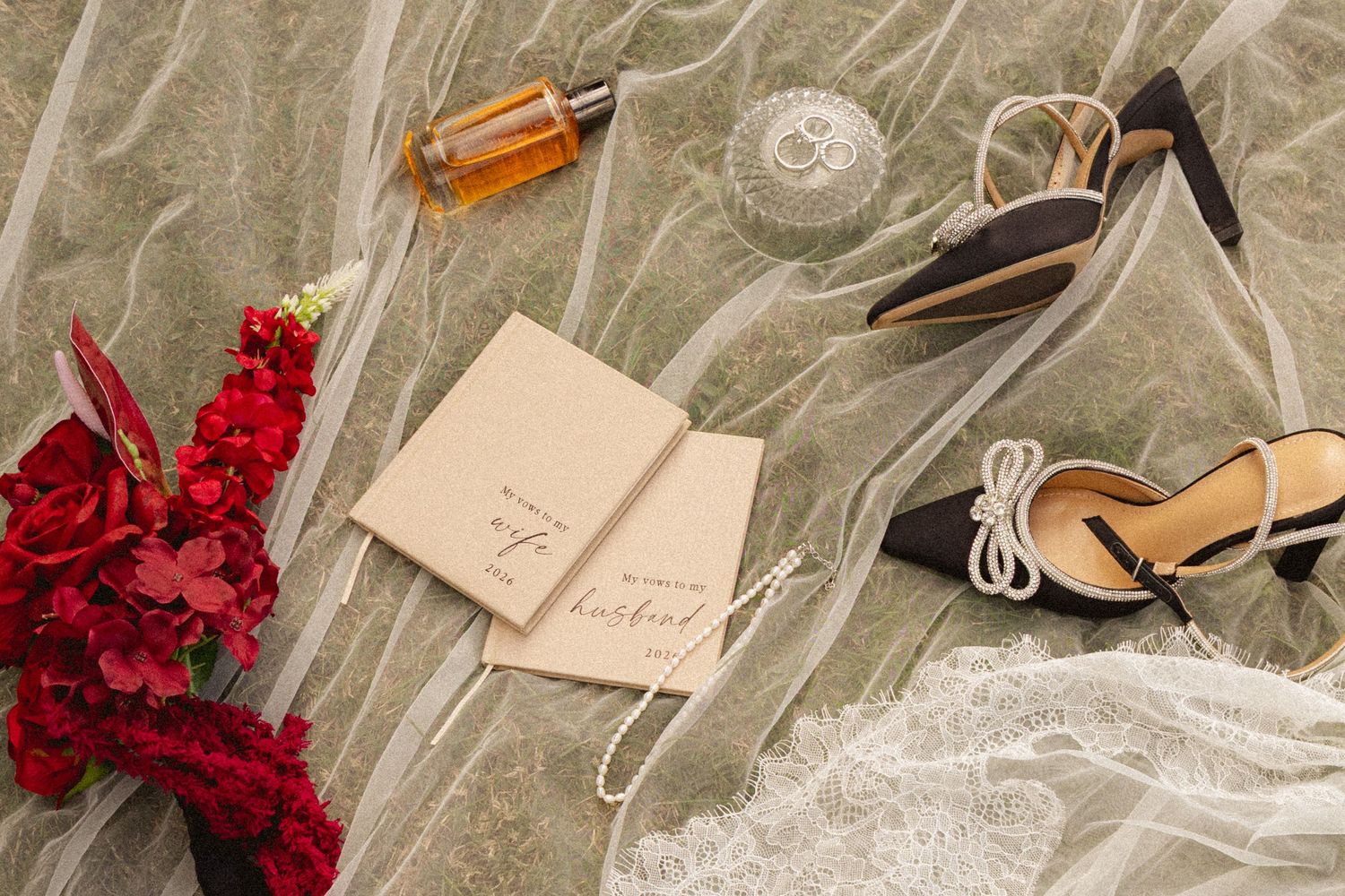 Elegant wedding flat lay with red florals, heels, veil, perfume, and handwritten vow books on sage green fabric.