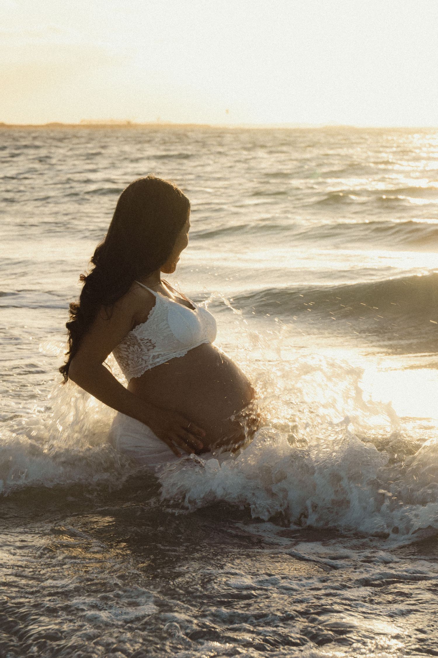 Pregnant woman in white lace bikini cradling baby bump in ocean waves at golden sunset on beach.