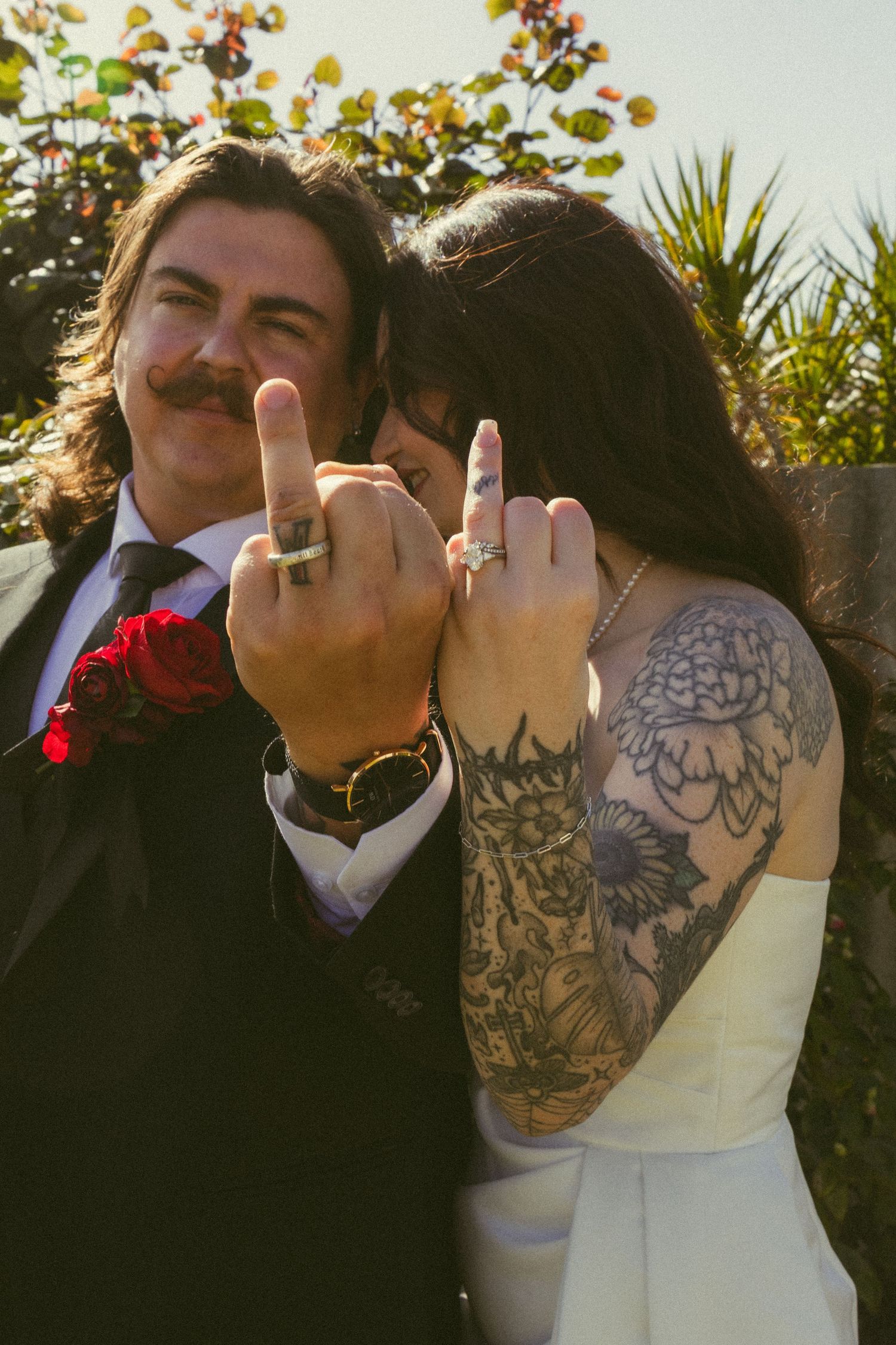 Newlyweds showing off wedding rings with middle fingers, groom in suit with mustache, bride with tattoos in white dress.