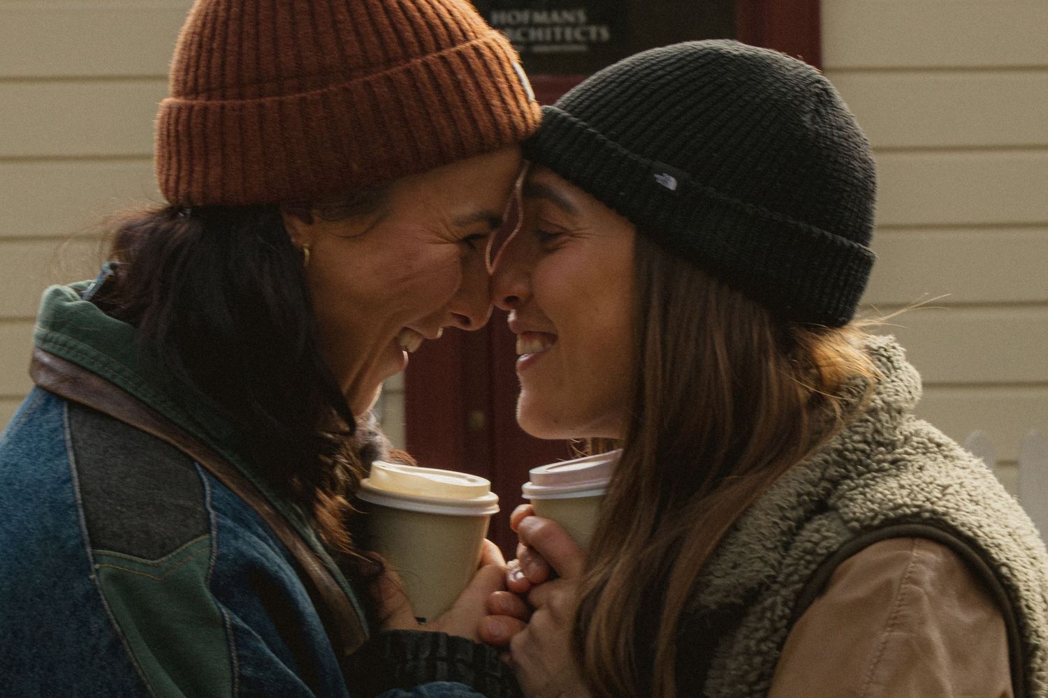Two women in winter beanies share a tender forehead-to-forehead moment while holding coffee cups outdoors.