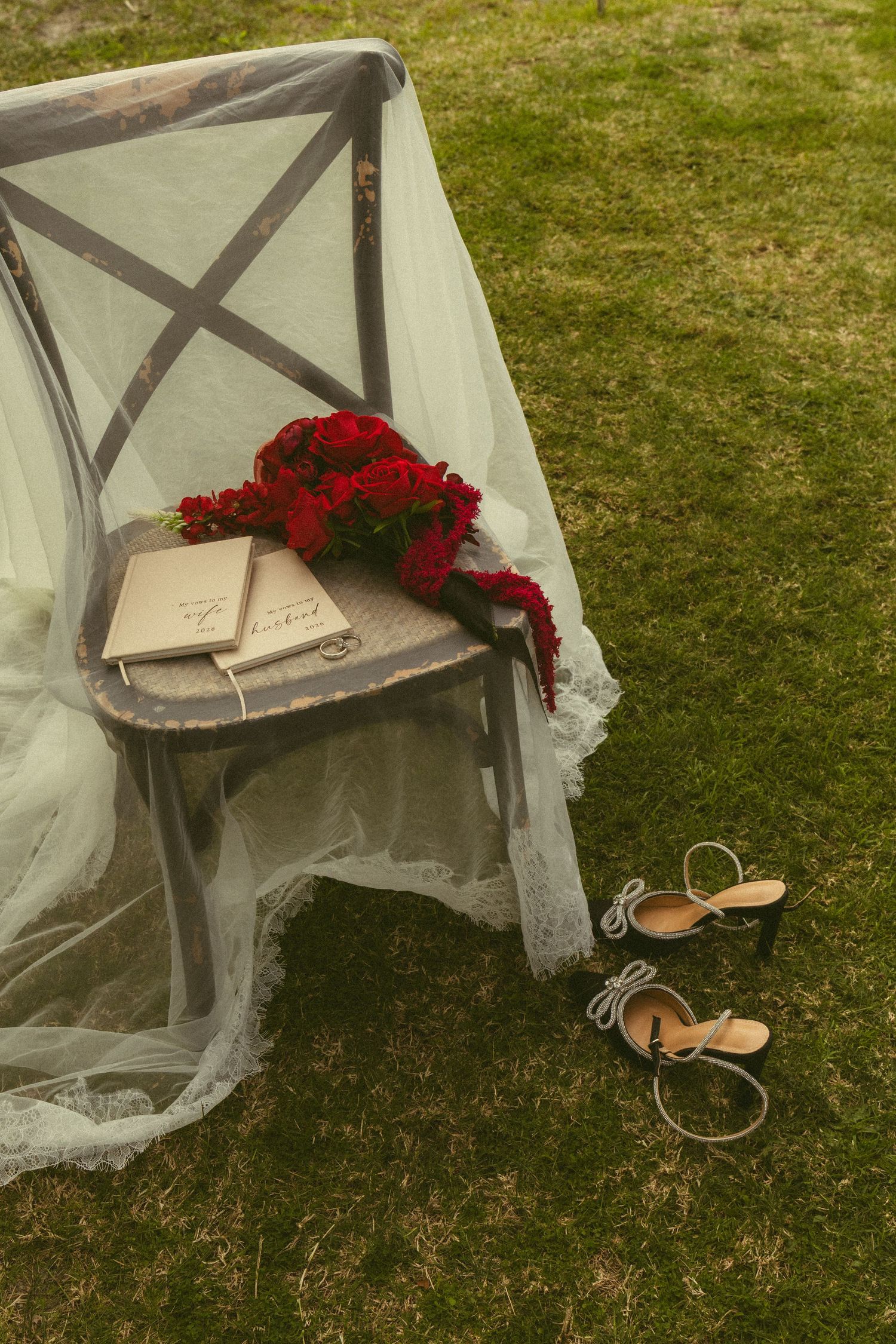 Wedding veil draped over chair with red rose bouquet, vows book and heels.