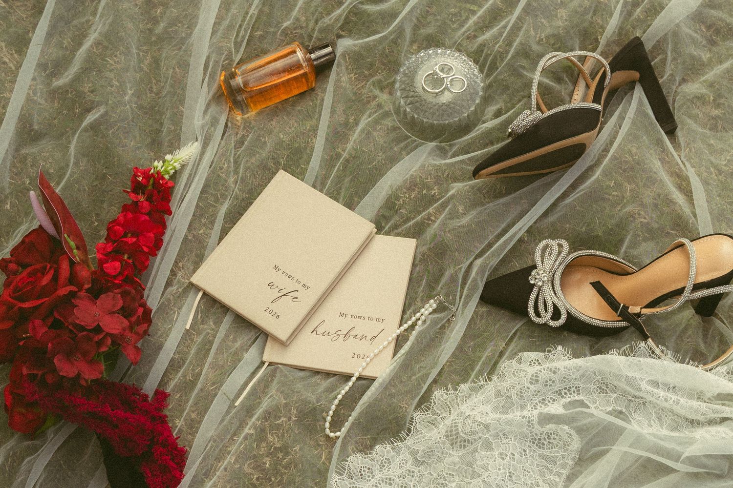 Wedding flat lay with veil, red florals, vow books, perfume and heeled sandals.