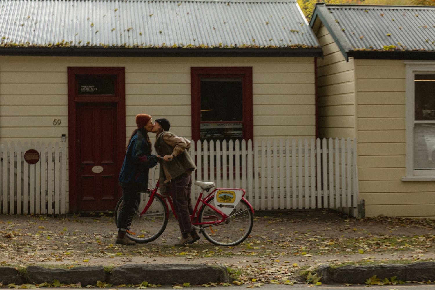 Couple kissing beside a red bicycle in front of a white picket fence and vintage cottage.