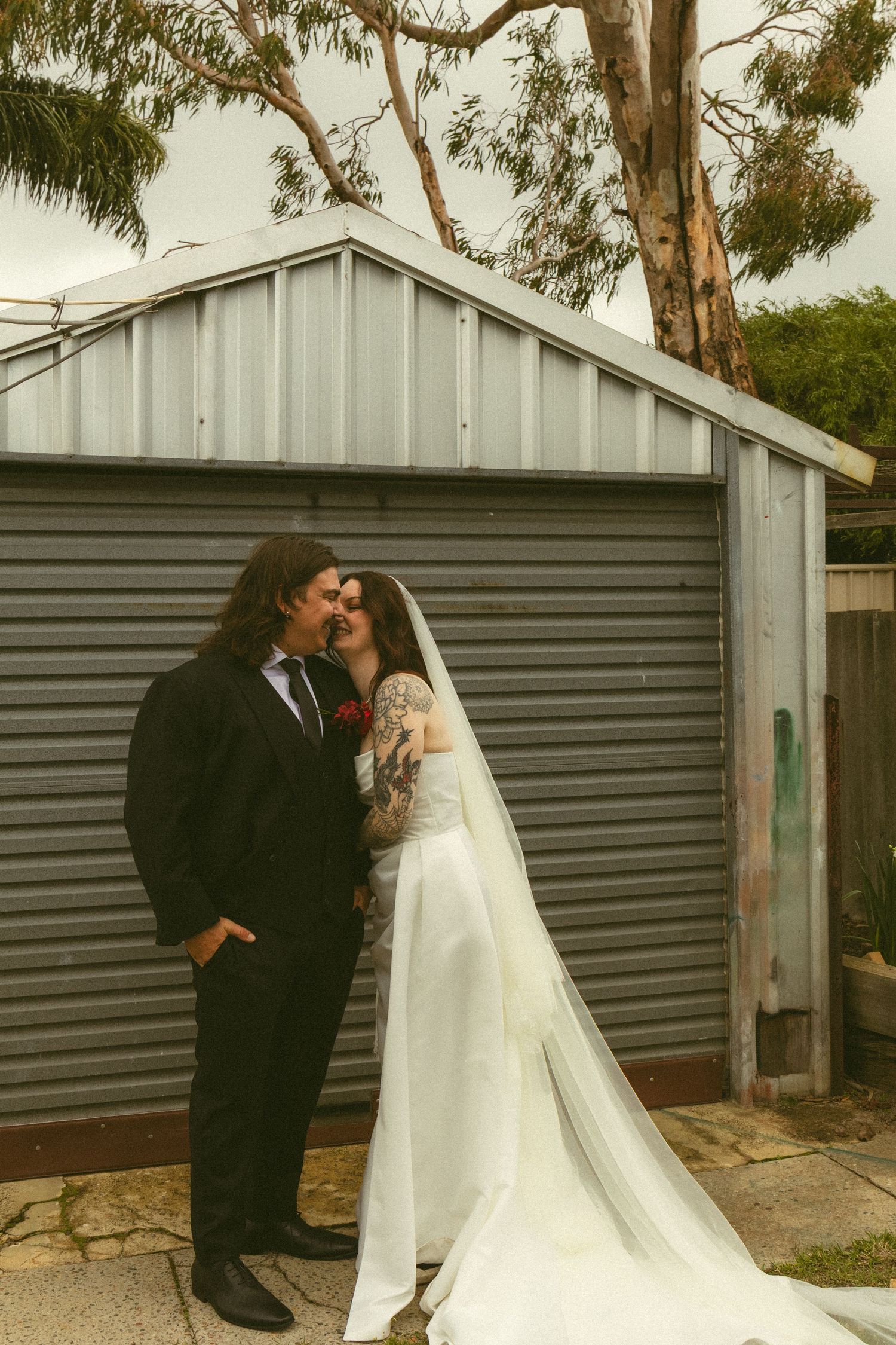 Bride and groom share a romantic kiss in wedding attire outside a rustic corrugated iron shed with eucalyptus tree.