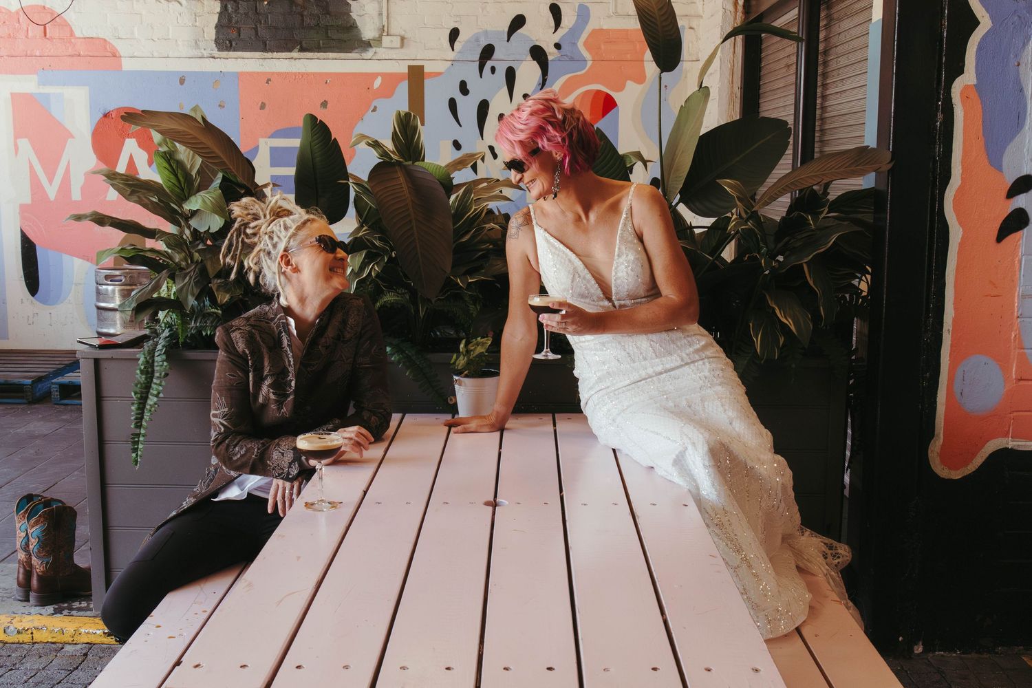 Bride in white lace gown with pink hair sits at pastel table chatting with guest amid colorful mural backdrop.