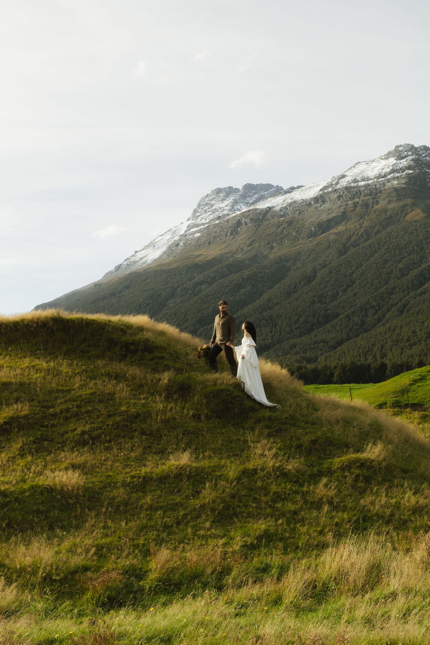 Wedding couple holds hands on green hillside with snow-capped mountain backdrop.