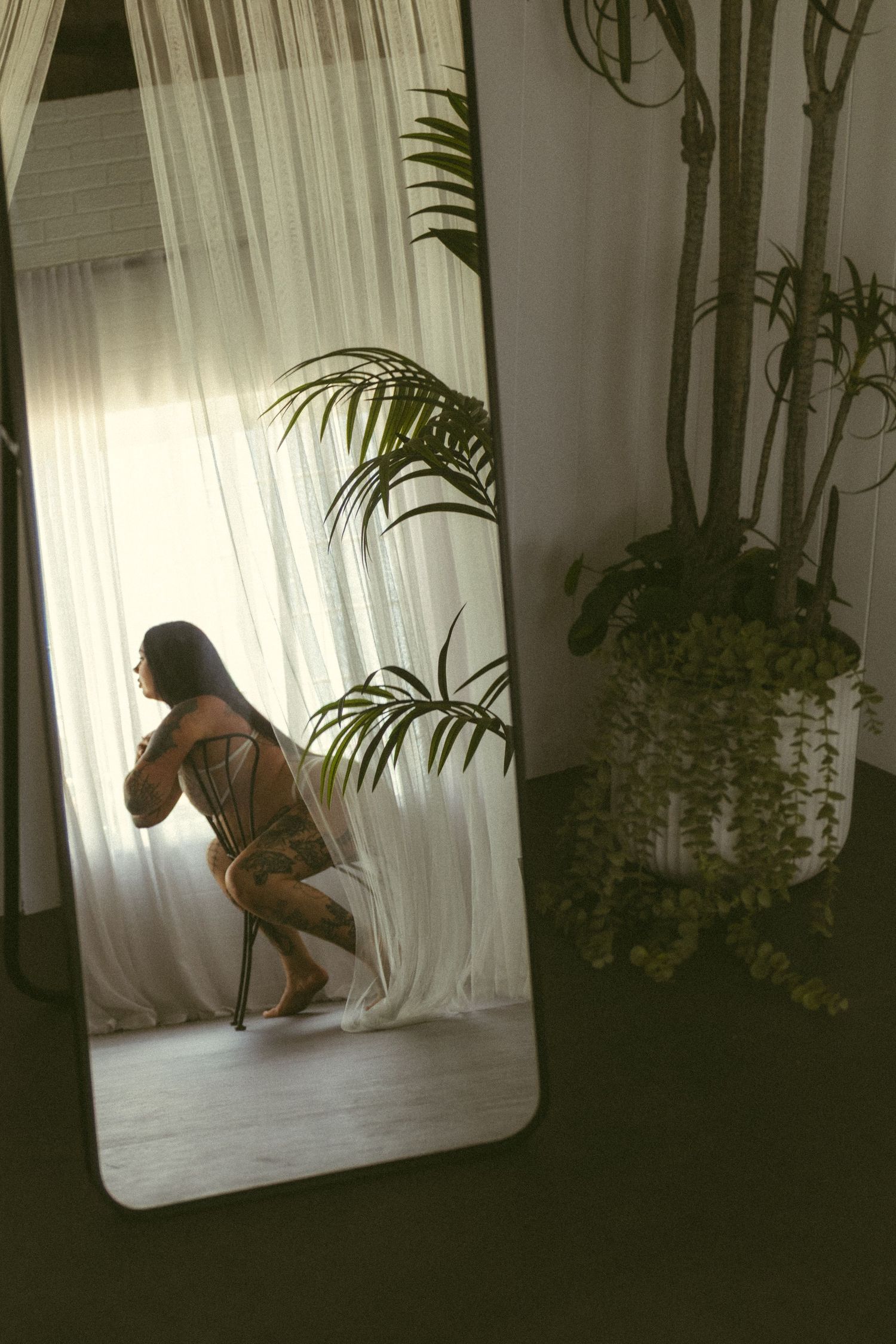 Woman in lingerie posing by sheer curtains with tropical plants in a softly lit room mirror reflection.