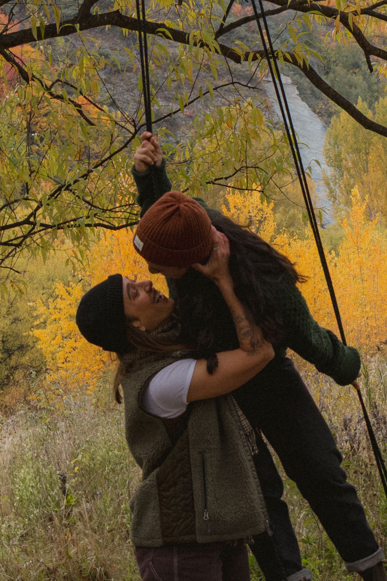 Couple sharing a romantic moment under autumn tree branches with vibrant yellow fall foliage and river in background.