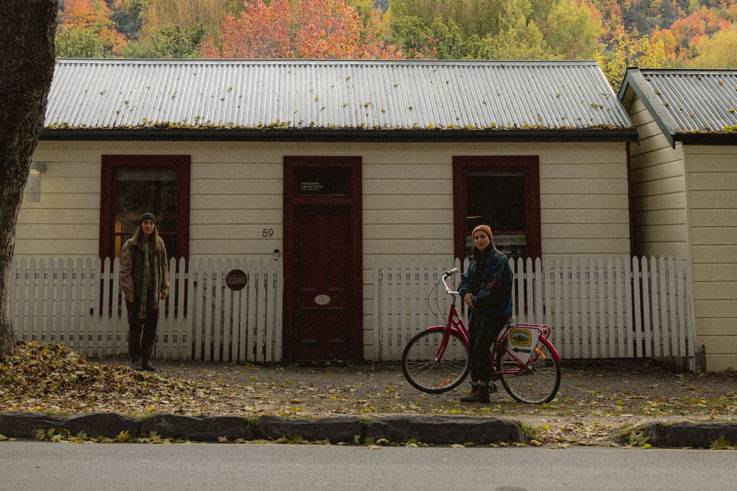 Two people stand outside a white cottage with red doors, surrounded by autumn foliage, one holding a red bicycle.