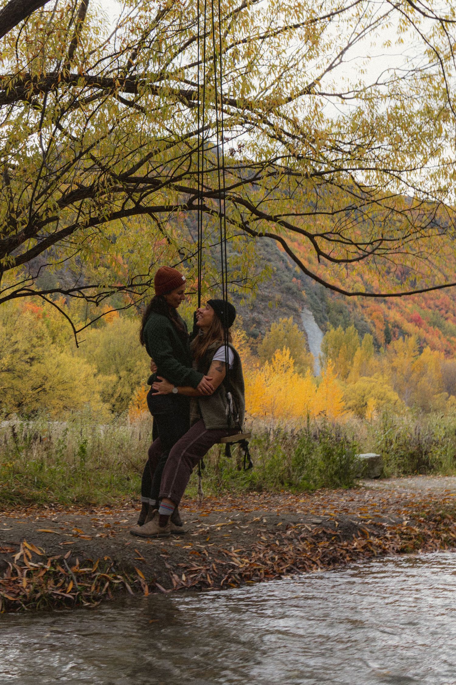 Couple sharing a romantic moment on a tree swing by a river surrounded by vibrant autumn foliage.