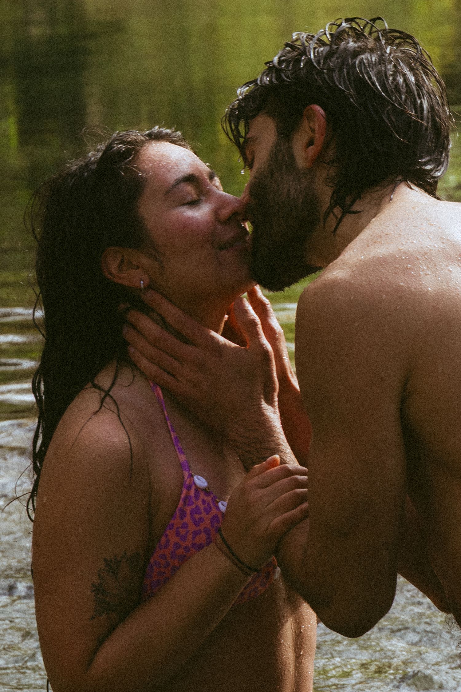 A couple sharing a romantic kiss in a natural water setting, with wet hair and summer swimwear.
