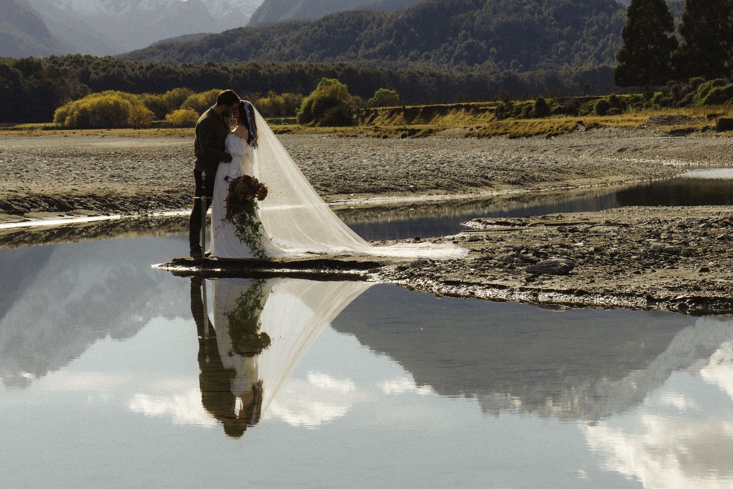 Bride and groom kissing near a reflective mountain lake, with wedding veil flowing, creating a stunning mirror image reflection.