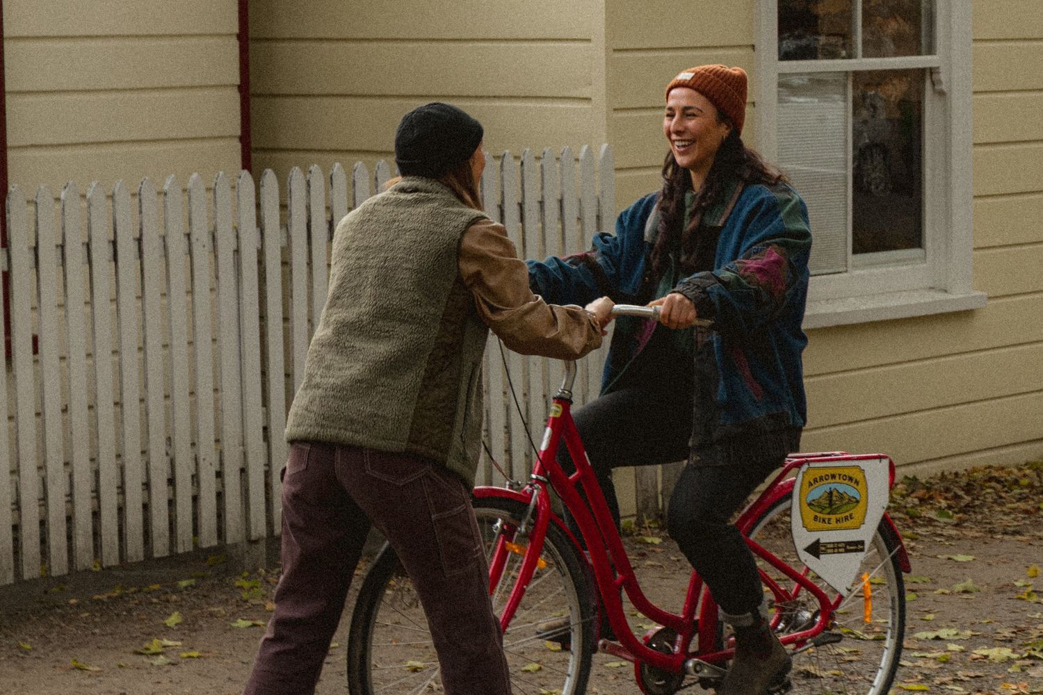 Two people sharing a joyful moment outdoors, one riding a red bicycle near a white picket fence.