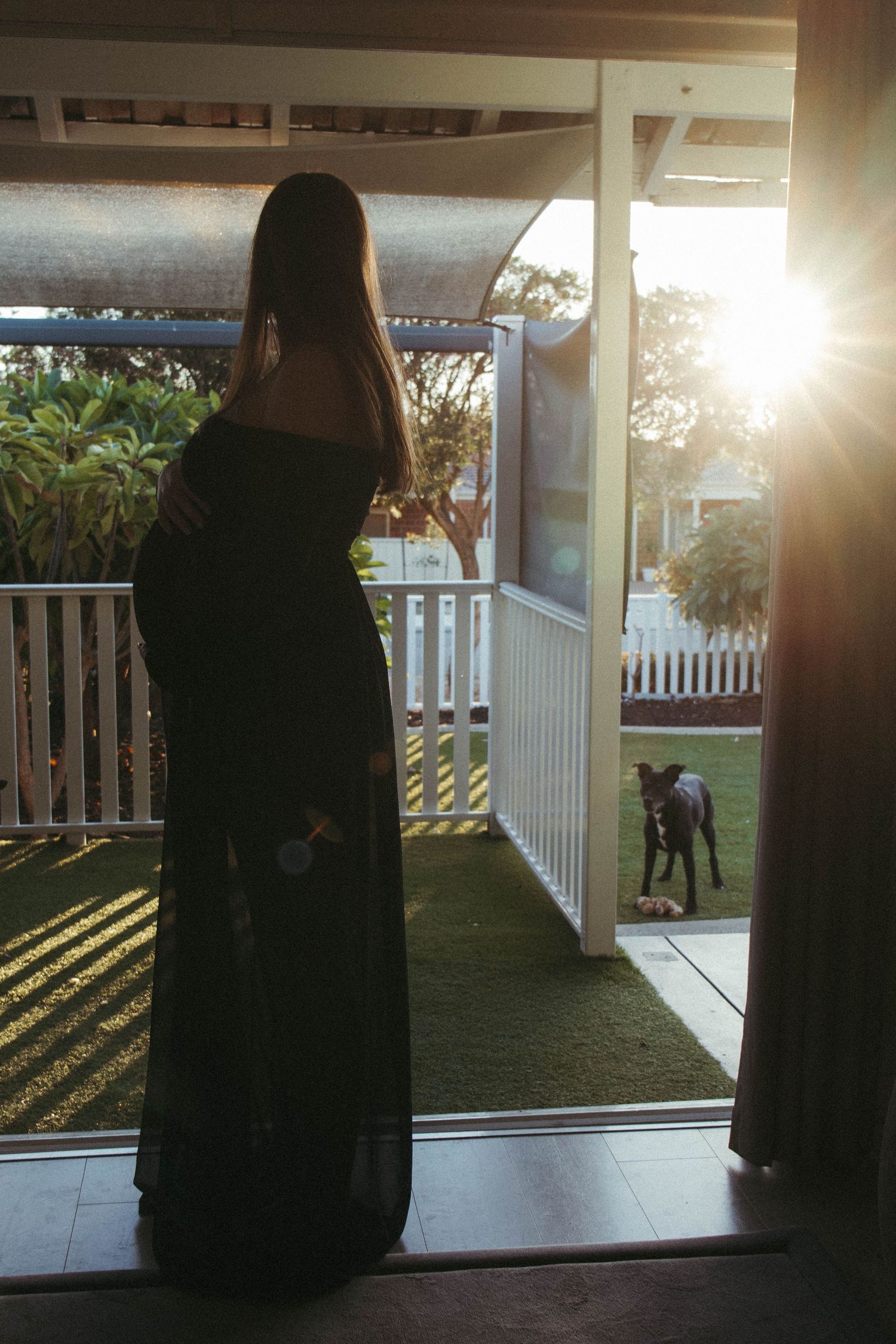 Pregnant woman in long dark dress standing in doorway at golden hour with dog on porch in background.