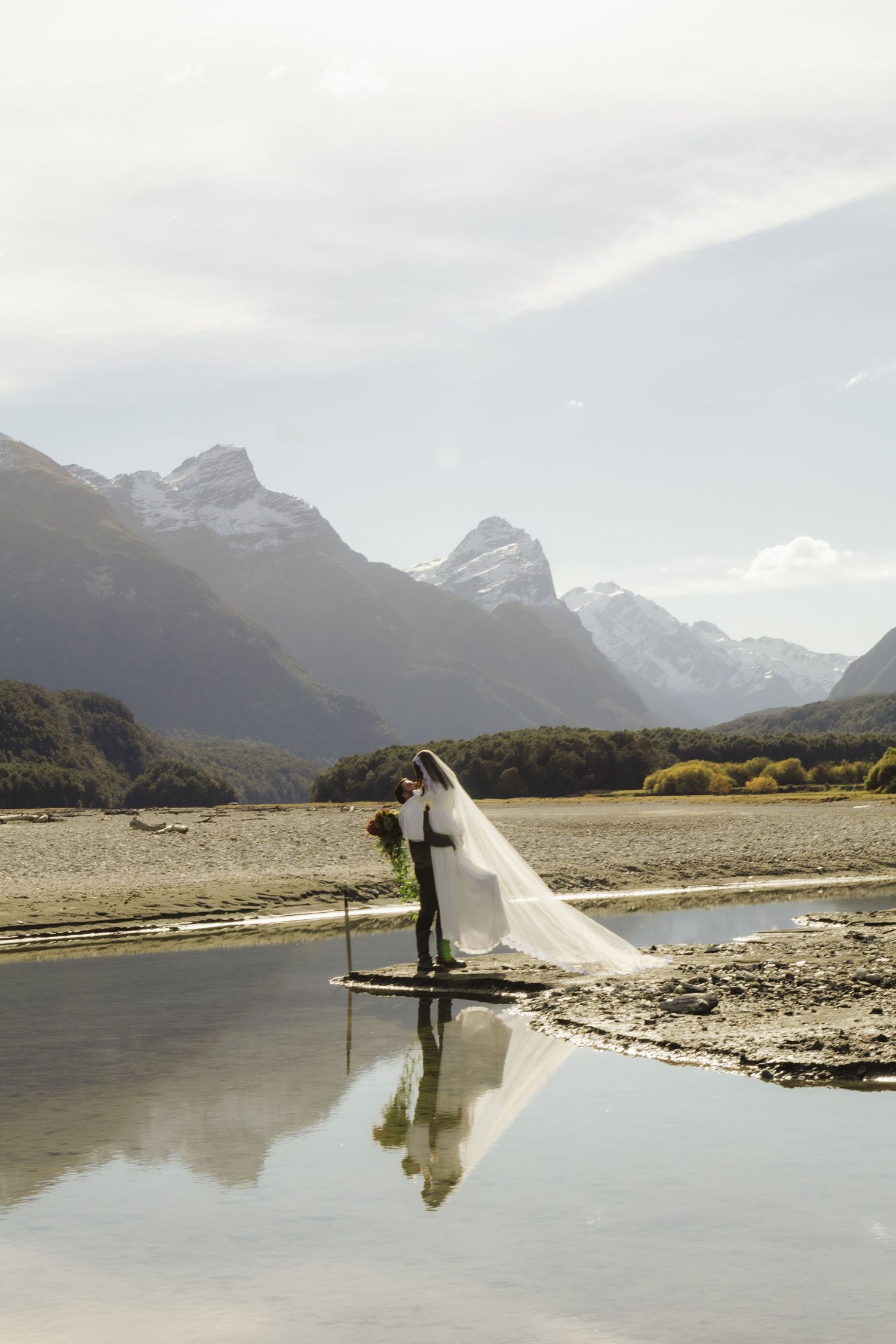 Bride and groom share a kiss reflected in still water with snow-capped New Zealand mountains backdrop.