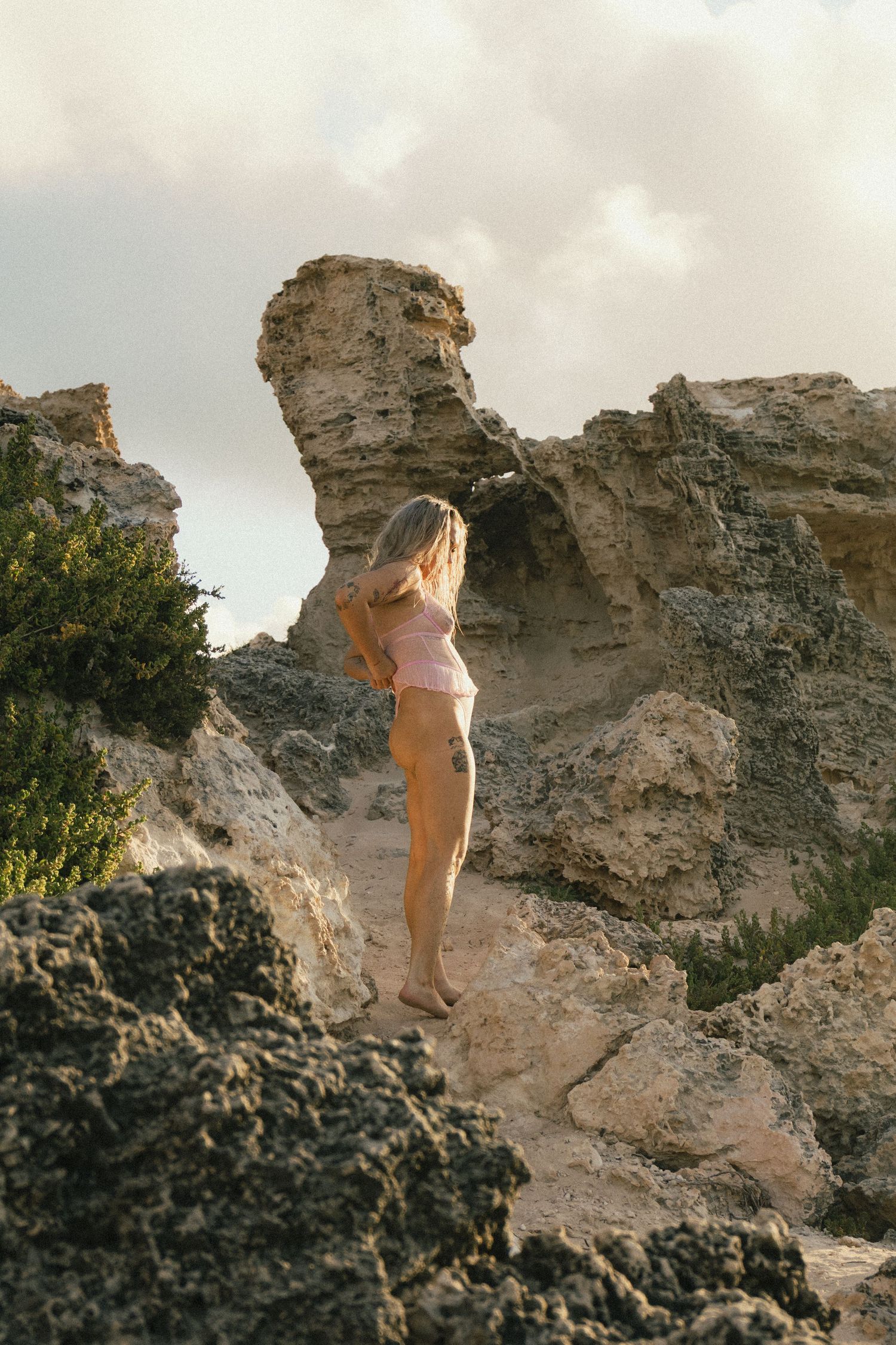 Woman in pink bikini standing on rocky coastal cliffs with dramatic limestone formations under cloudy sky.