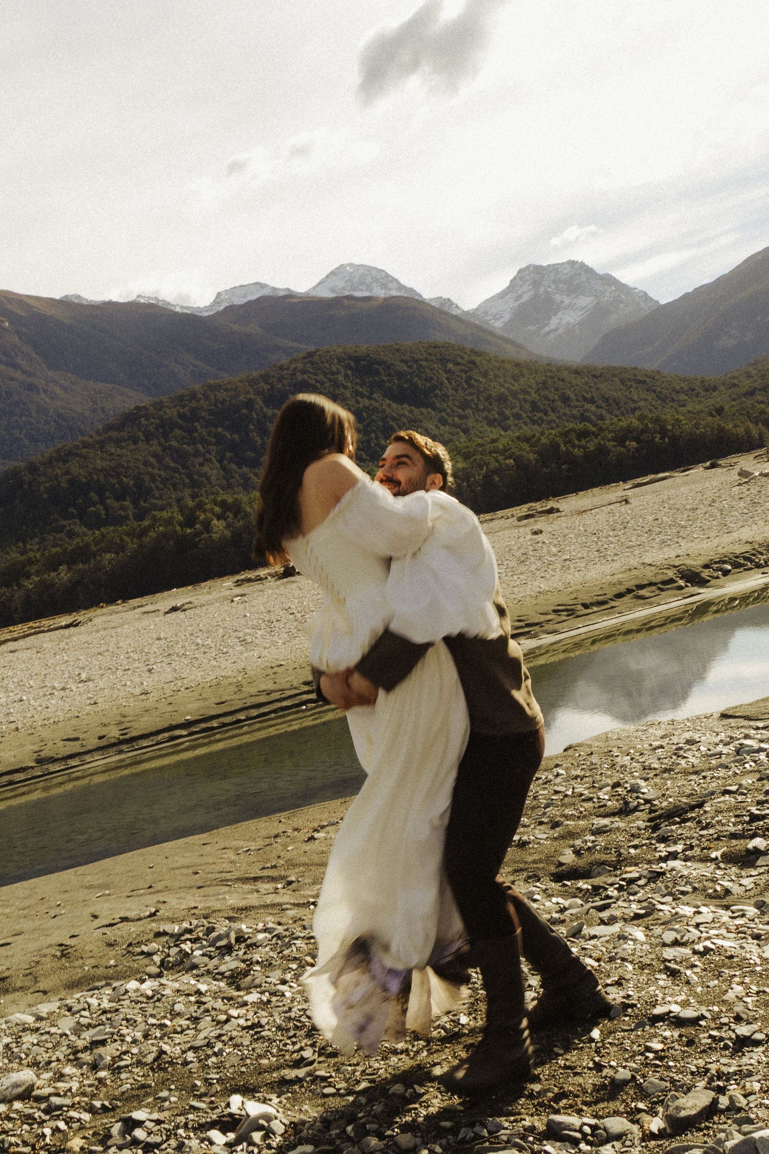 Couple embracing in wedding attire with stunning mountain backdrop near a rocky riverbank.