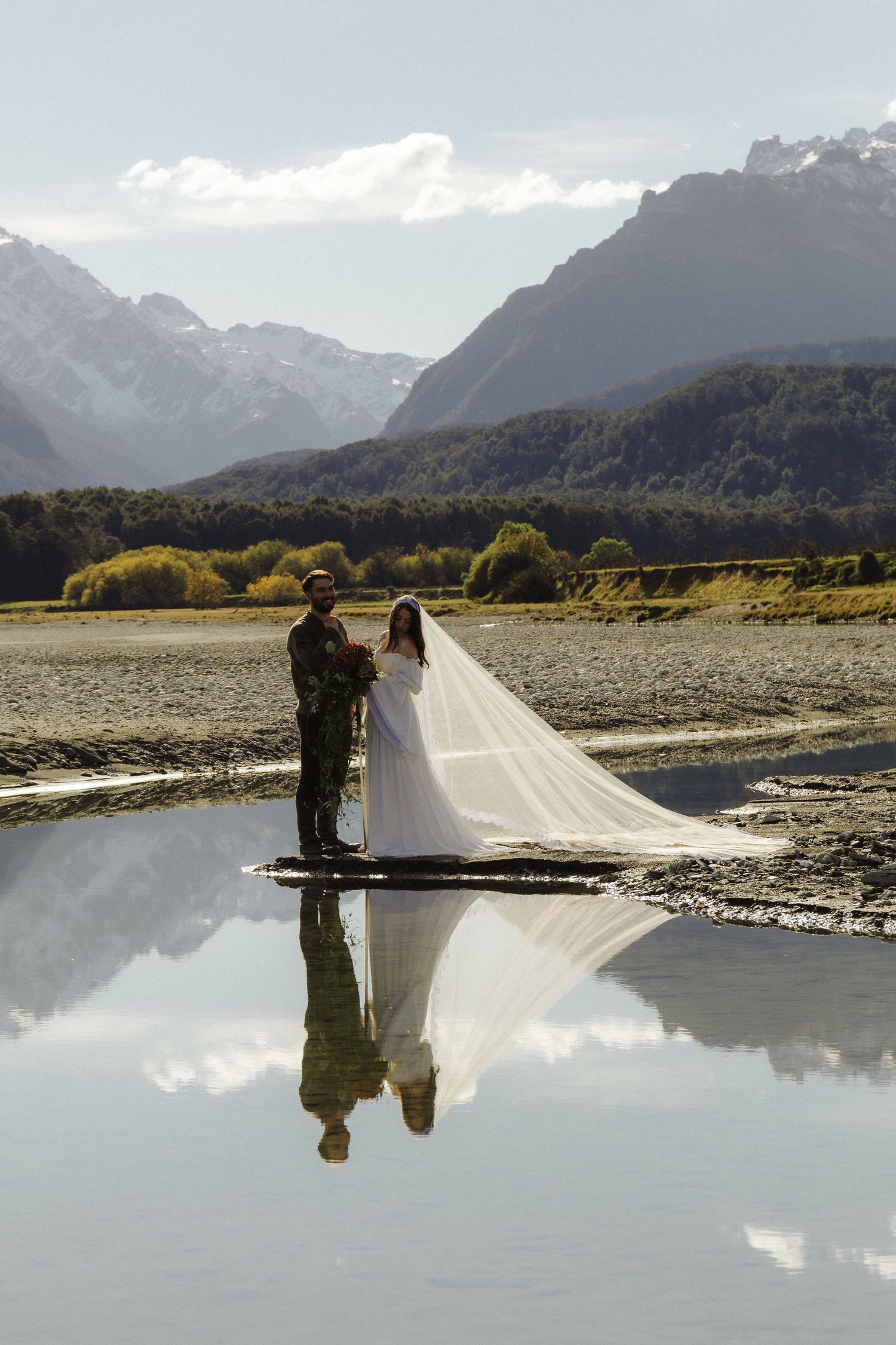 Bride and groom stand by reflective mountain lake, wedding veil flowing, snow-capped peaks backdrop, New Zealand elopement.