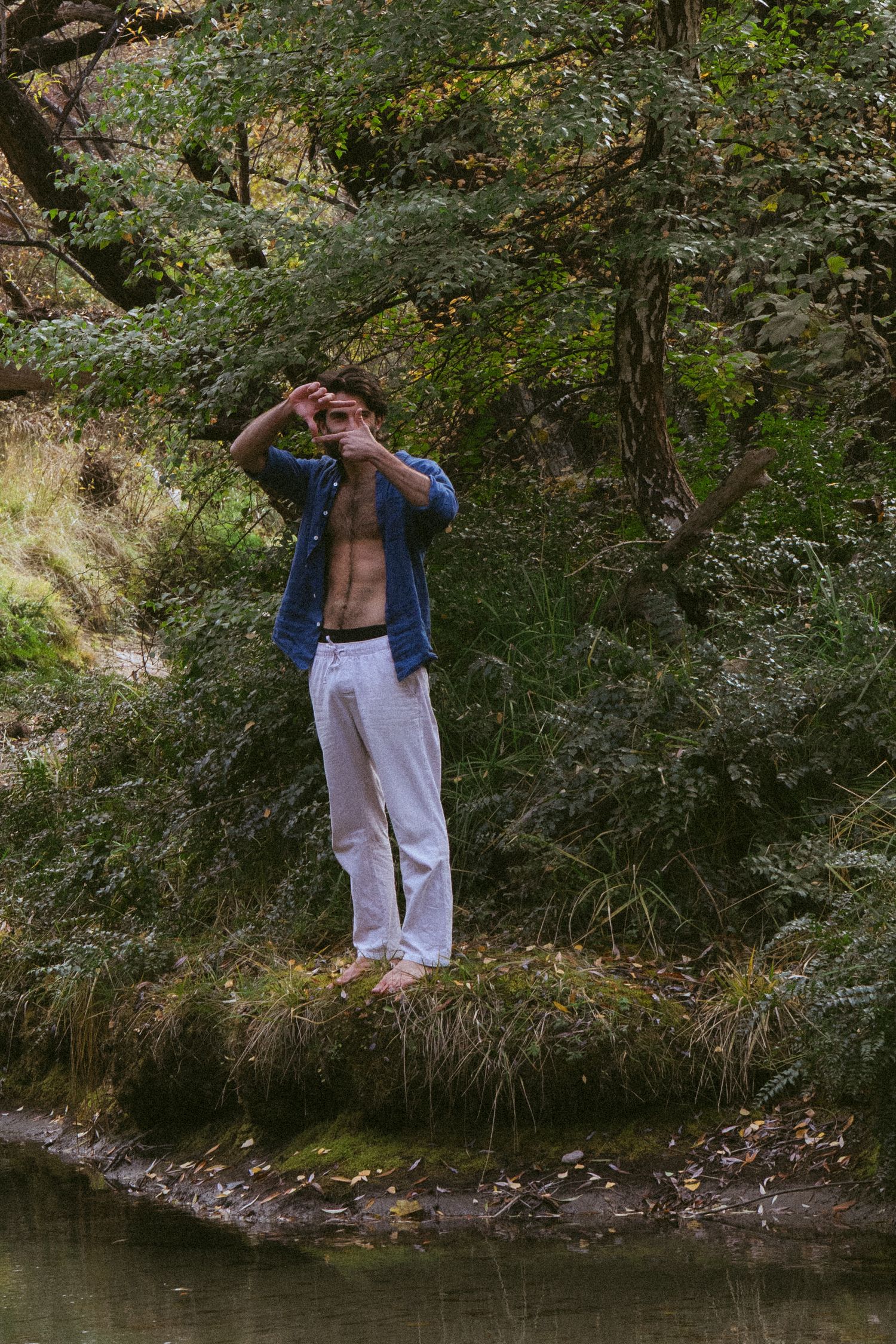 Shirtless man in white pants and open denim shirt standing barefoot by a forest stream surrounded by lush greenery.