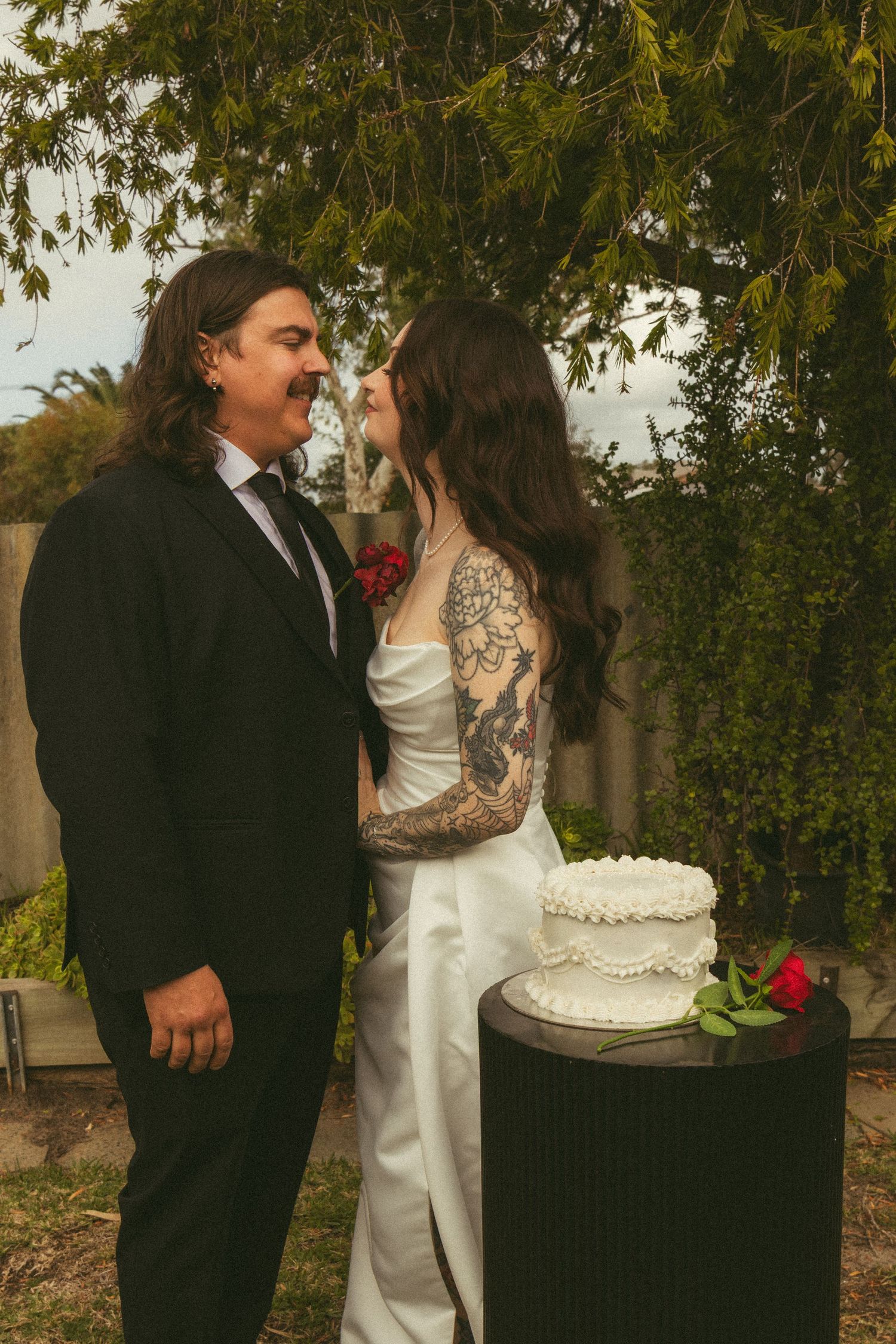 Bride with tattoos in white gown and groom in black suit stand by wedding cake outdoors under green trees.