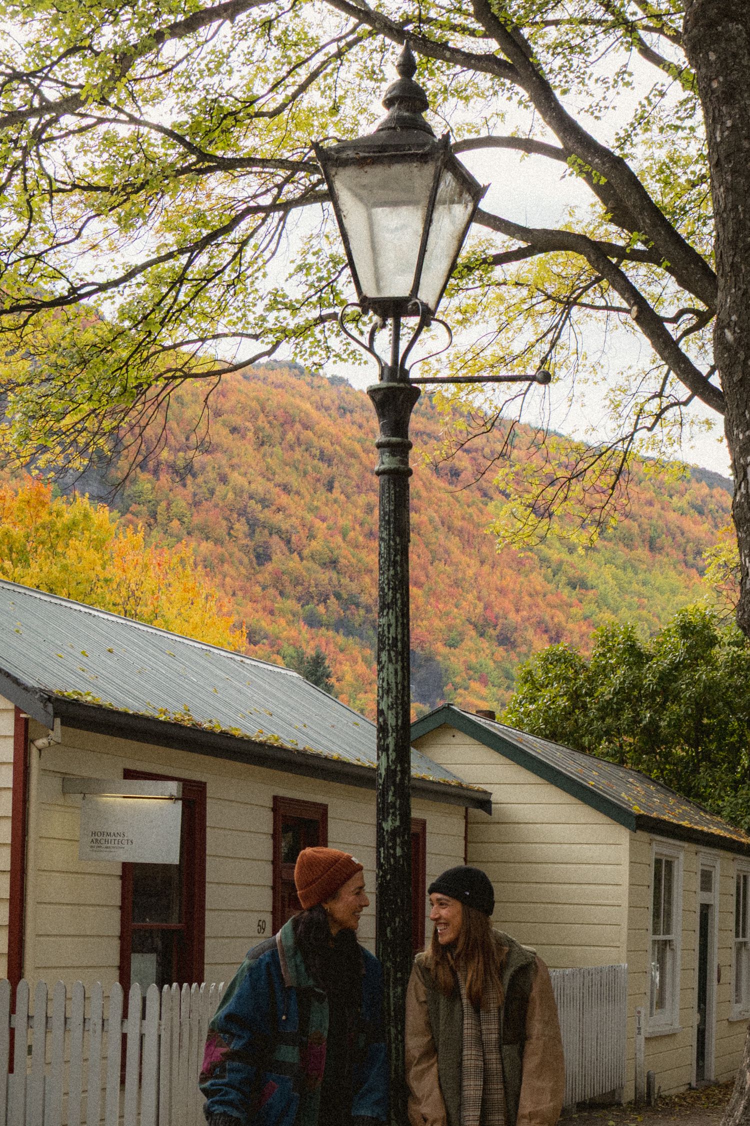 Two people chat by a vintage lamp post with colorful autumn foliage on a hillside behind quaint cottages.