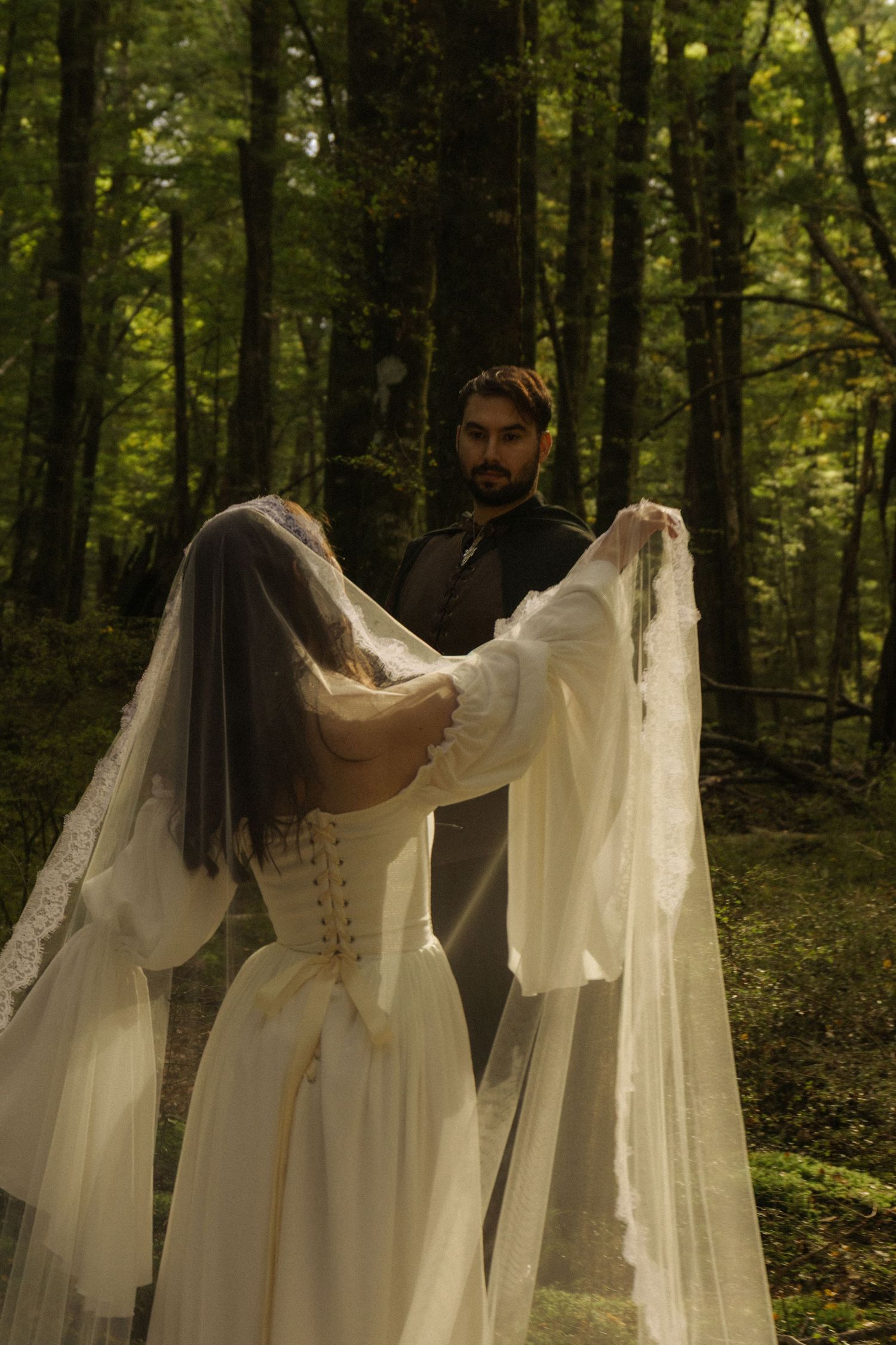 Bride in white corset gown and veil holds fabric toward bearded groom in dark vest during forest wedding ceremony.