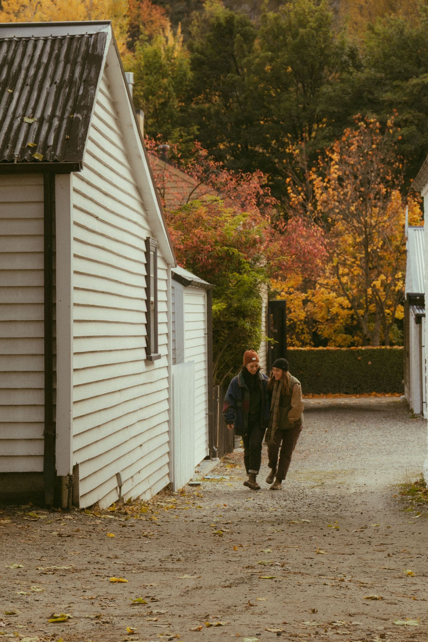 Two people walking between white wooden buildings on a gravel path surrounded by colorful autumn foliage.