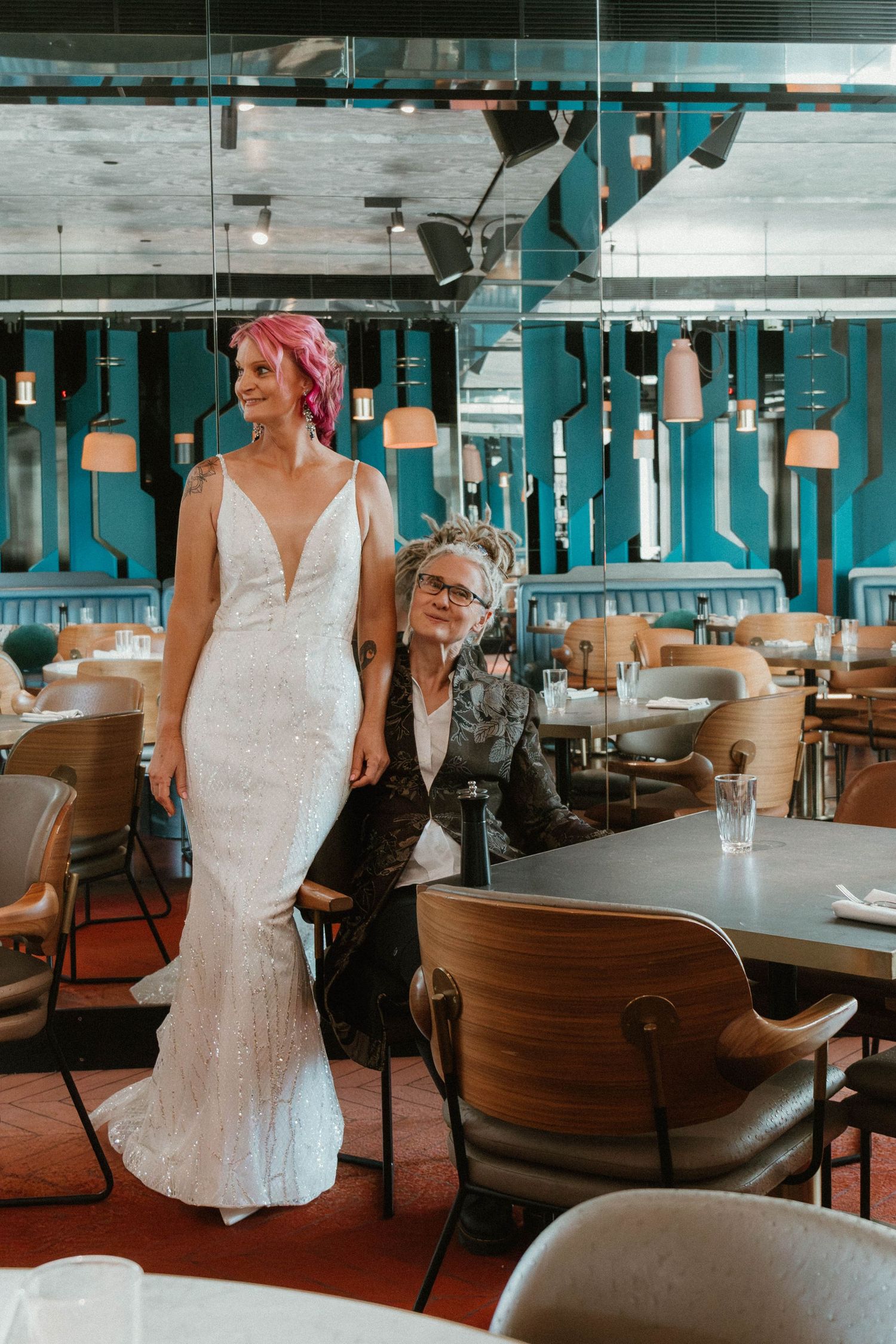 Bride in white dress with pink hair stands beside their seated wife in a visually chaotic room.