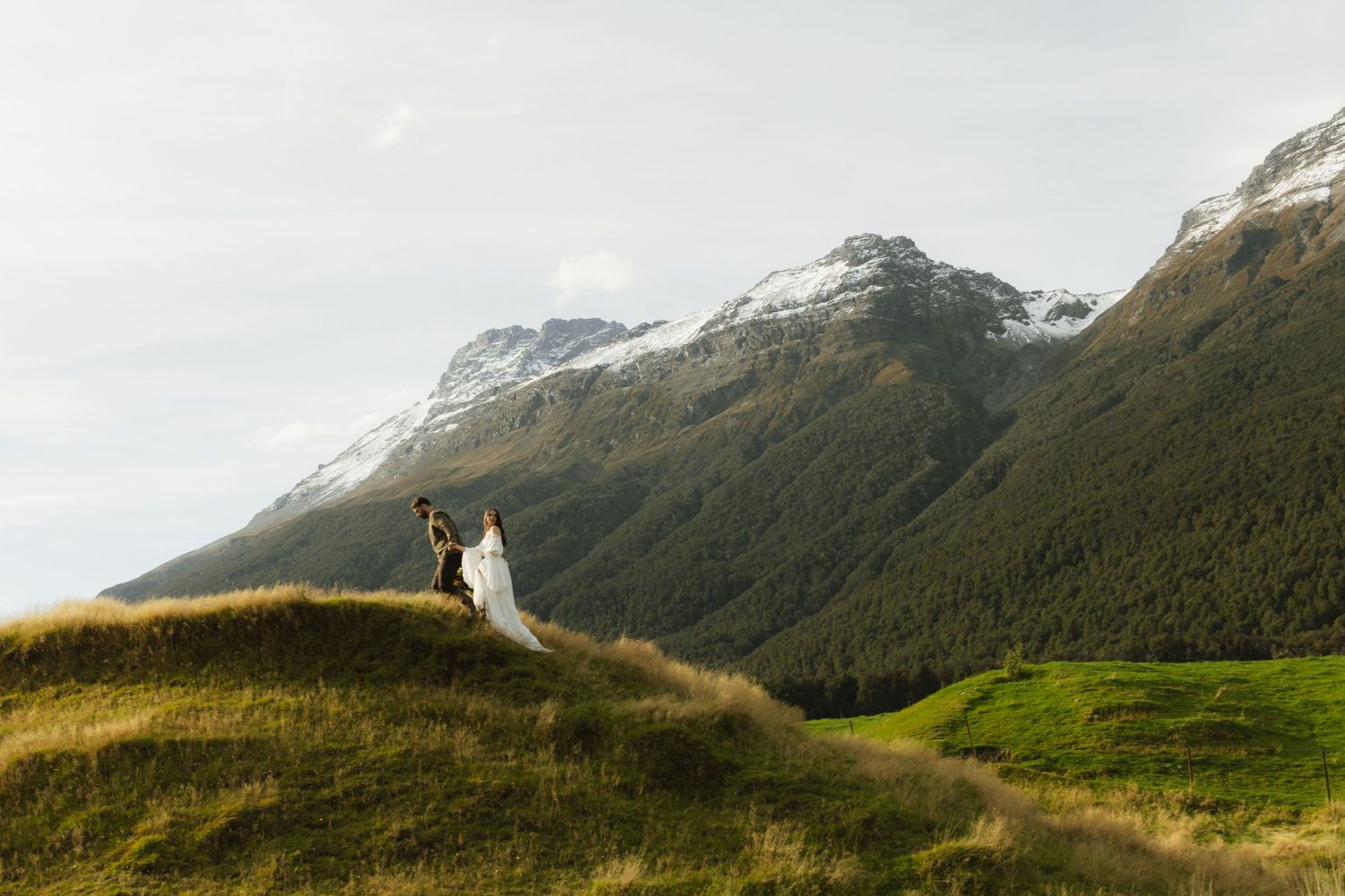 Bride and groom stand on a green hillside with snow-capped mountains in New Zealand backdrop.