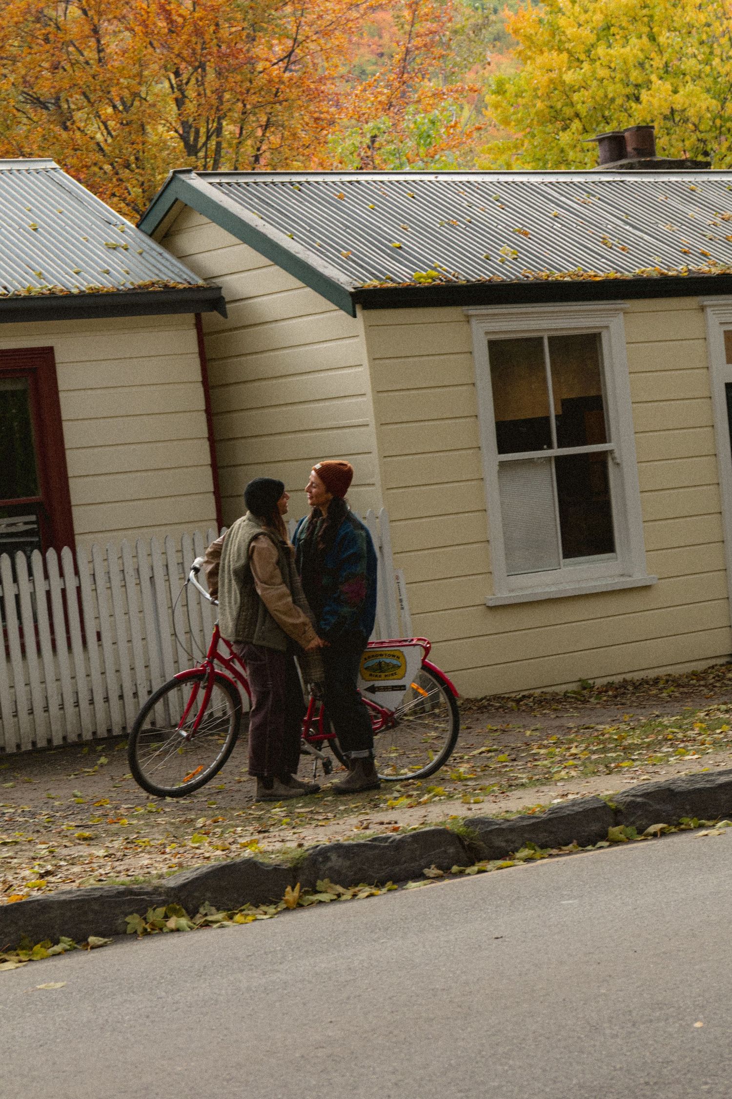 Couple sharing a romantic moment beside a red bicycle on an autumn street with colorful trees and a white cottage.