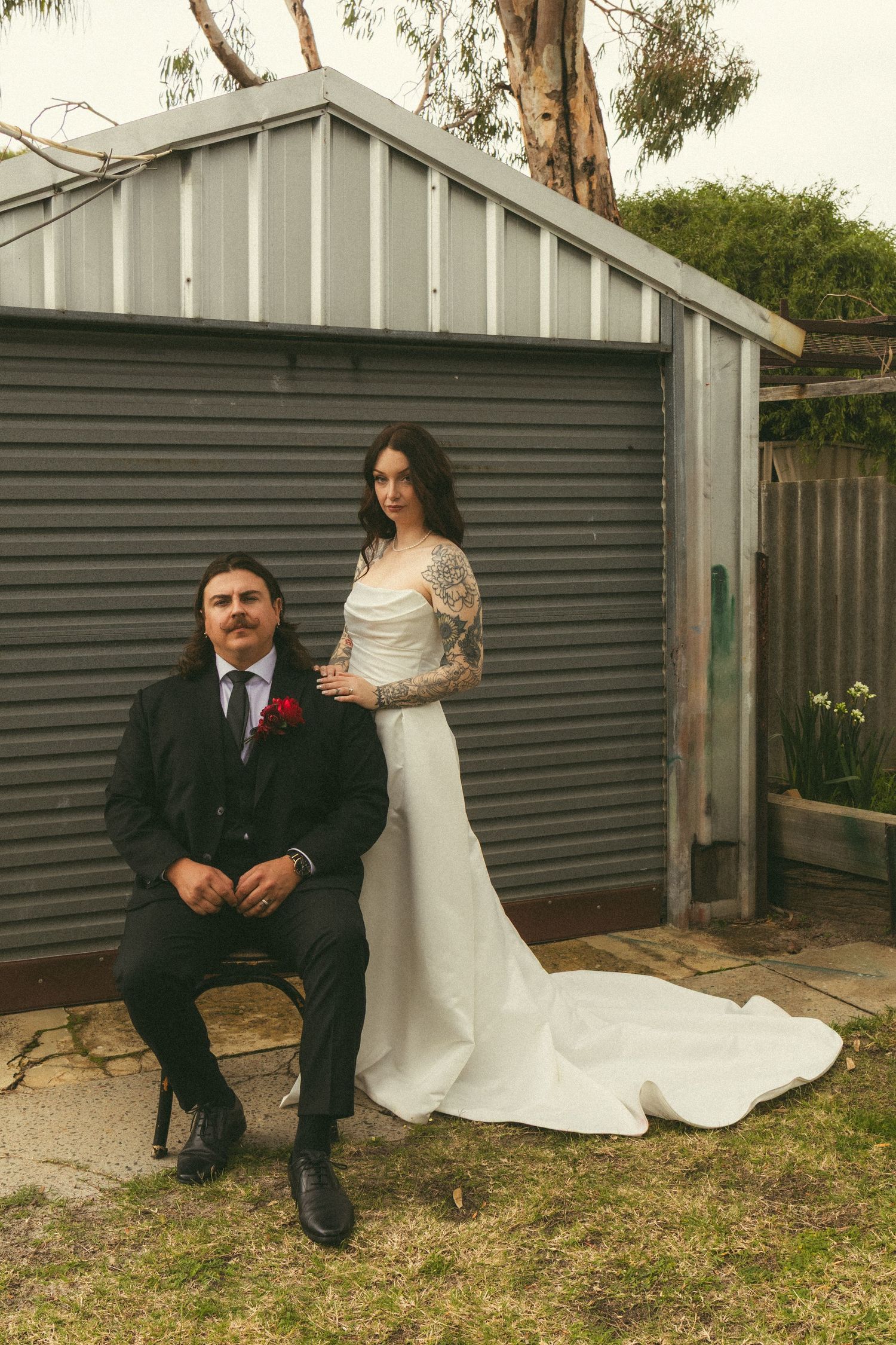 Bride in white gown with lace sleeves stands beside seated groom in black suit before gray shed backdrop.