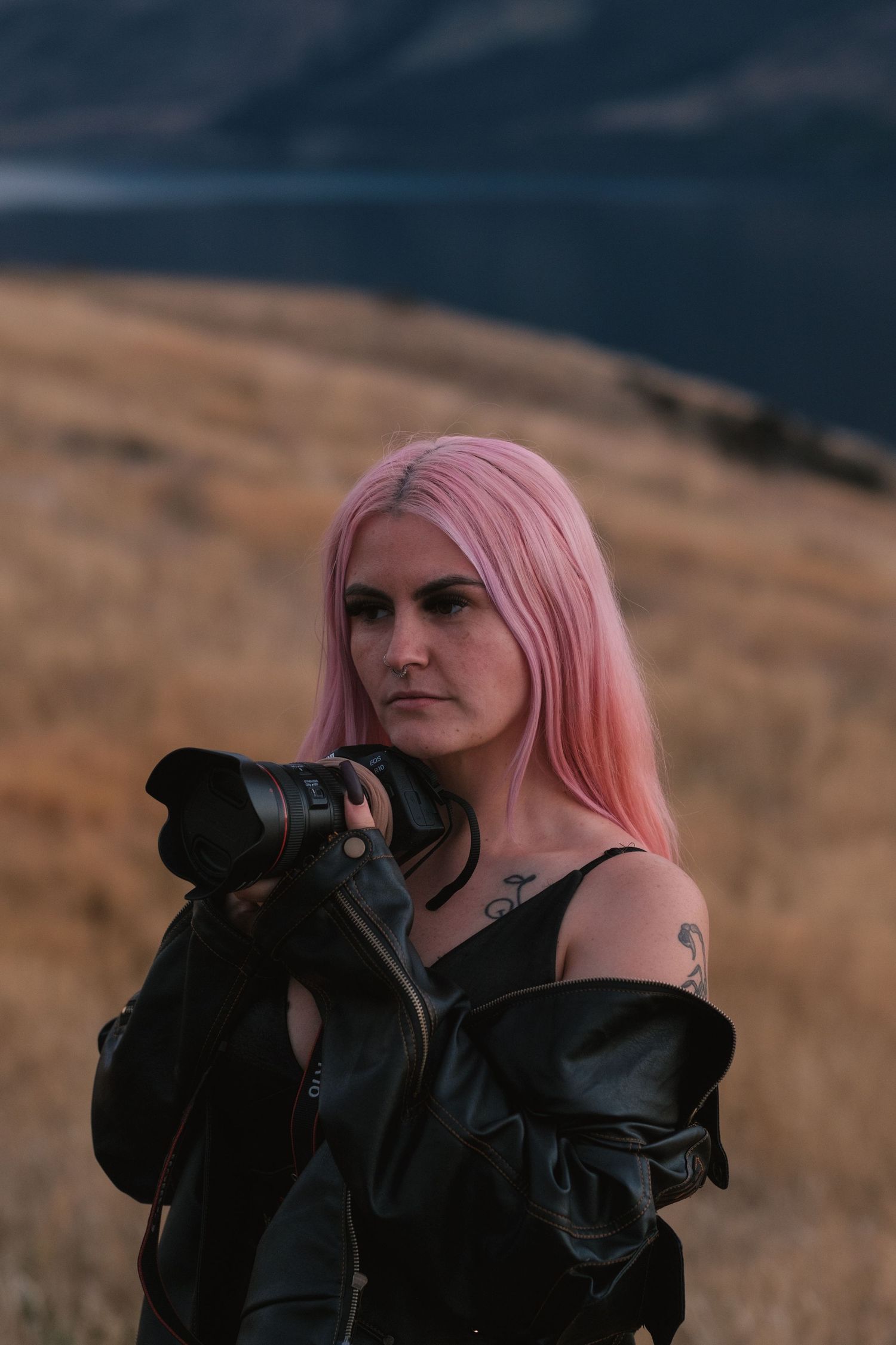 Woman with pink hair in black leather jacket holds camera outdoors near mountain landscape.