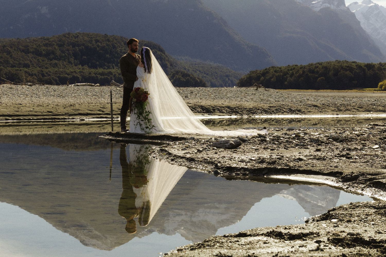 Bride and groom share a romantic kiss by a reflective mountain lake, with a flowing veil and stunning alpine scenery.