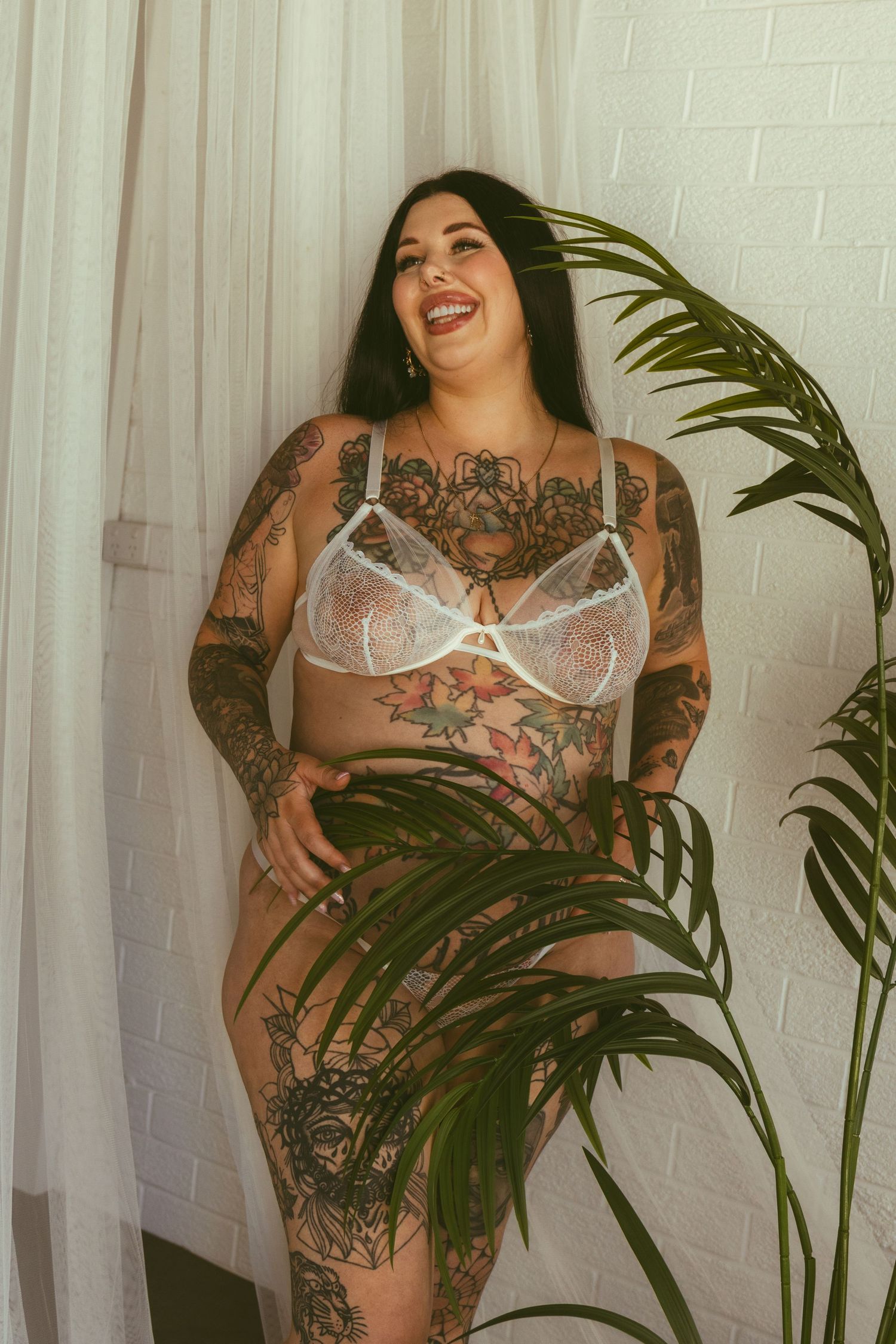 Heavily tattooed woman in white lace lingerie posing confidently beside a tropical palm plant indoors.