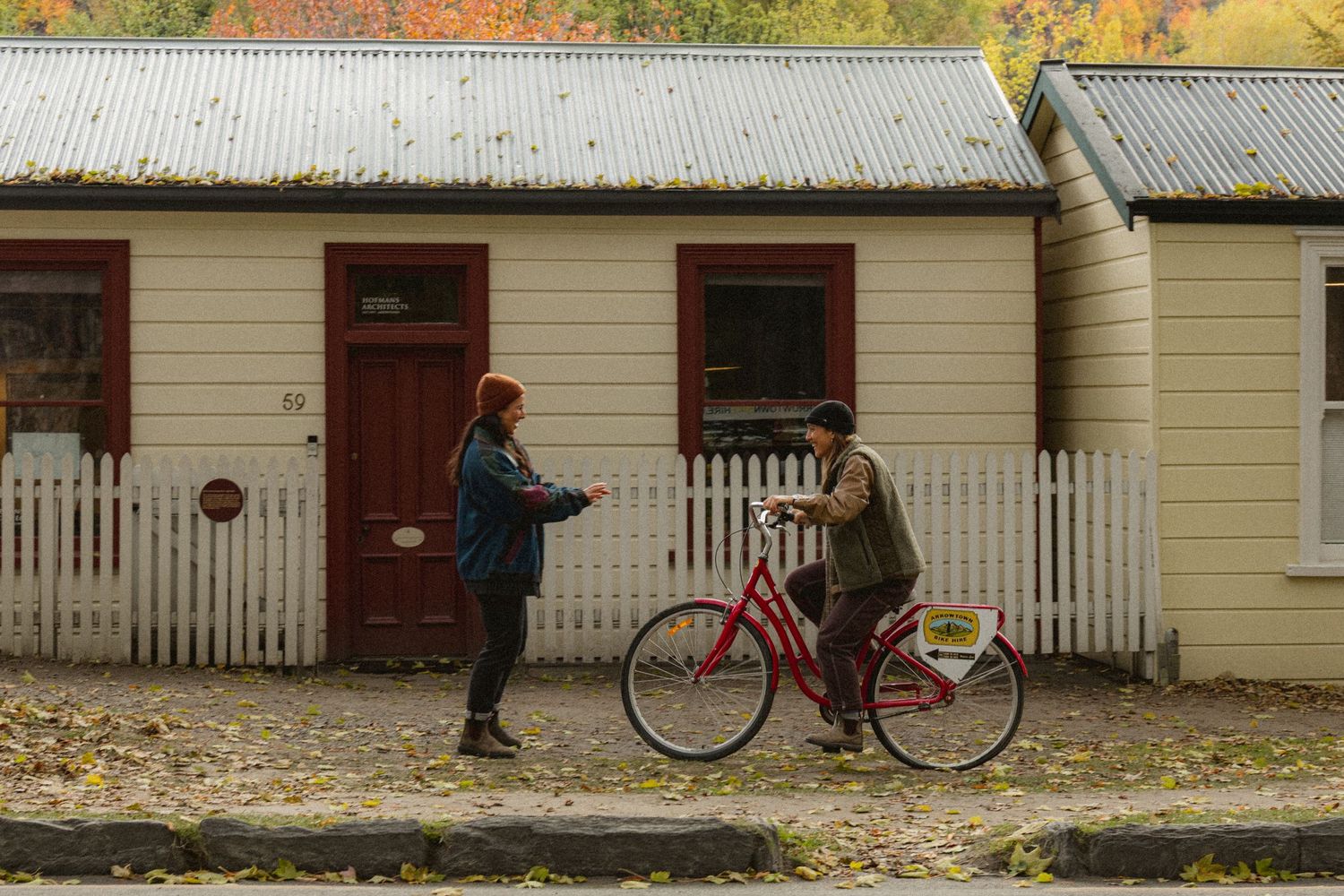 Two people chat outside a white picket fence cottage with a red bicycle, surrounded by autumn foliage.