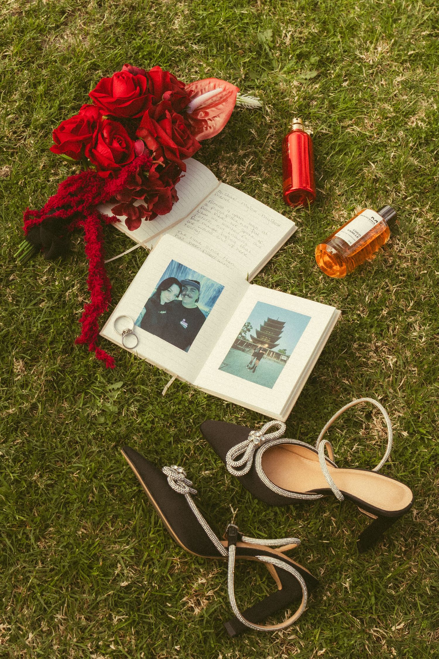 Flat lay of wedding accessories including red roses, Polaroids, perfume and heels on grass.