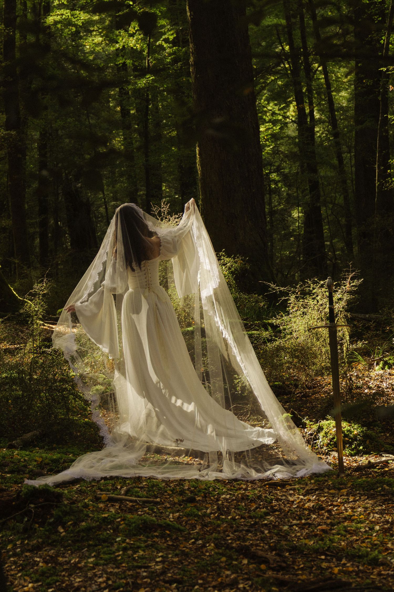 Bride in white wedding gown tosses flowing veil in a lush green forest.