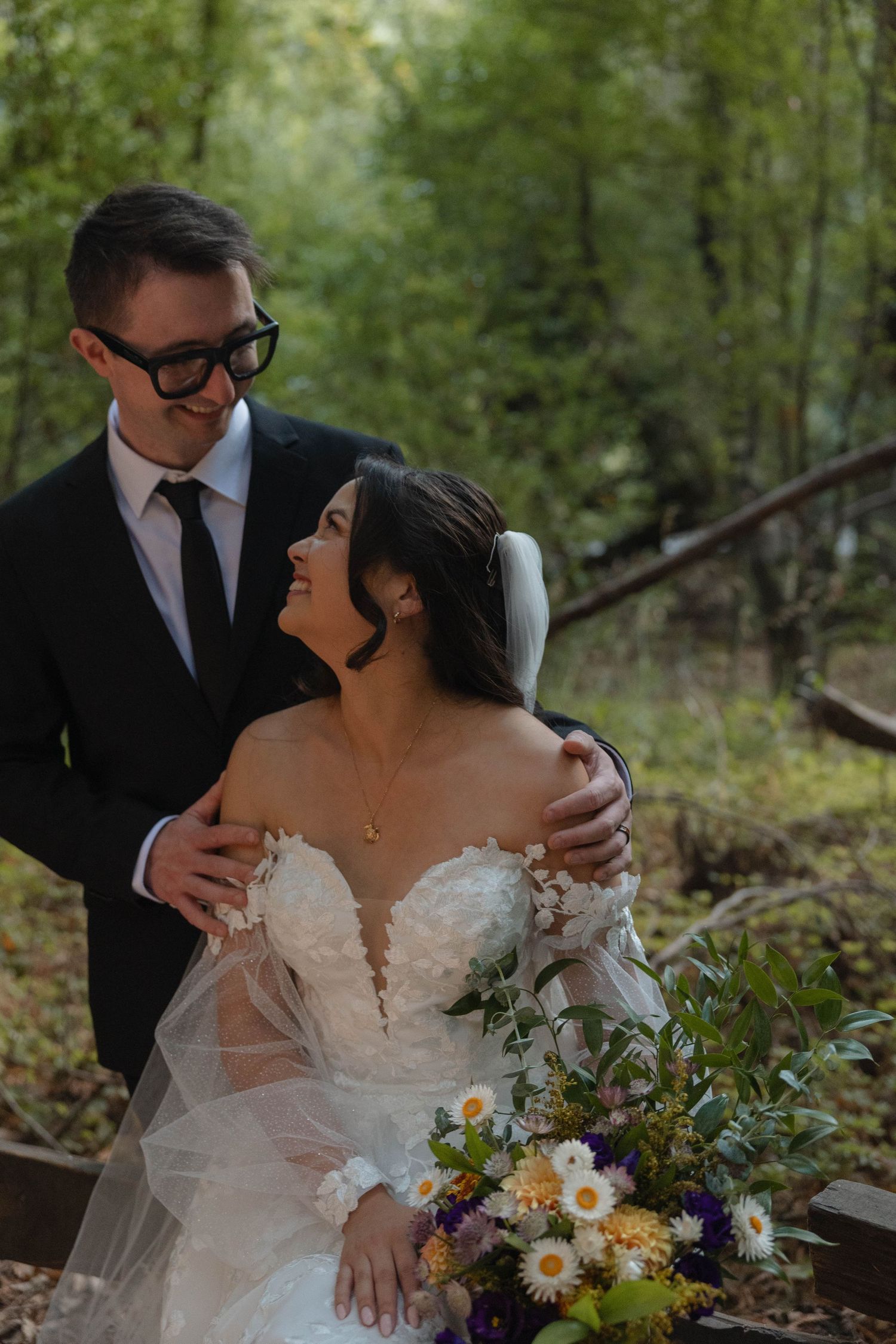 A groom in a black suit embraces a smiling bride in a floral gown holding a wildflower bouquet in a lush forest setting. Wedding photography