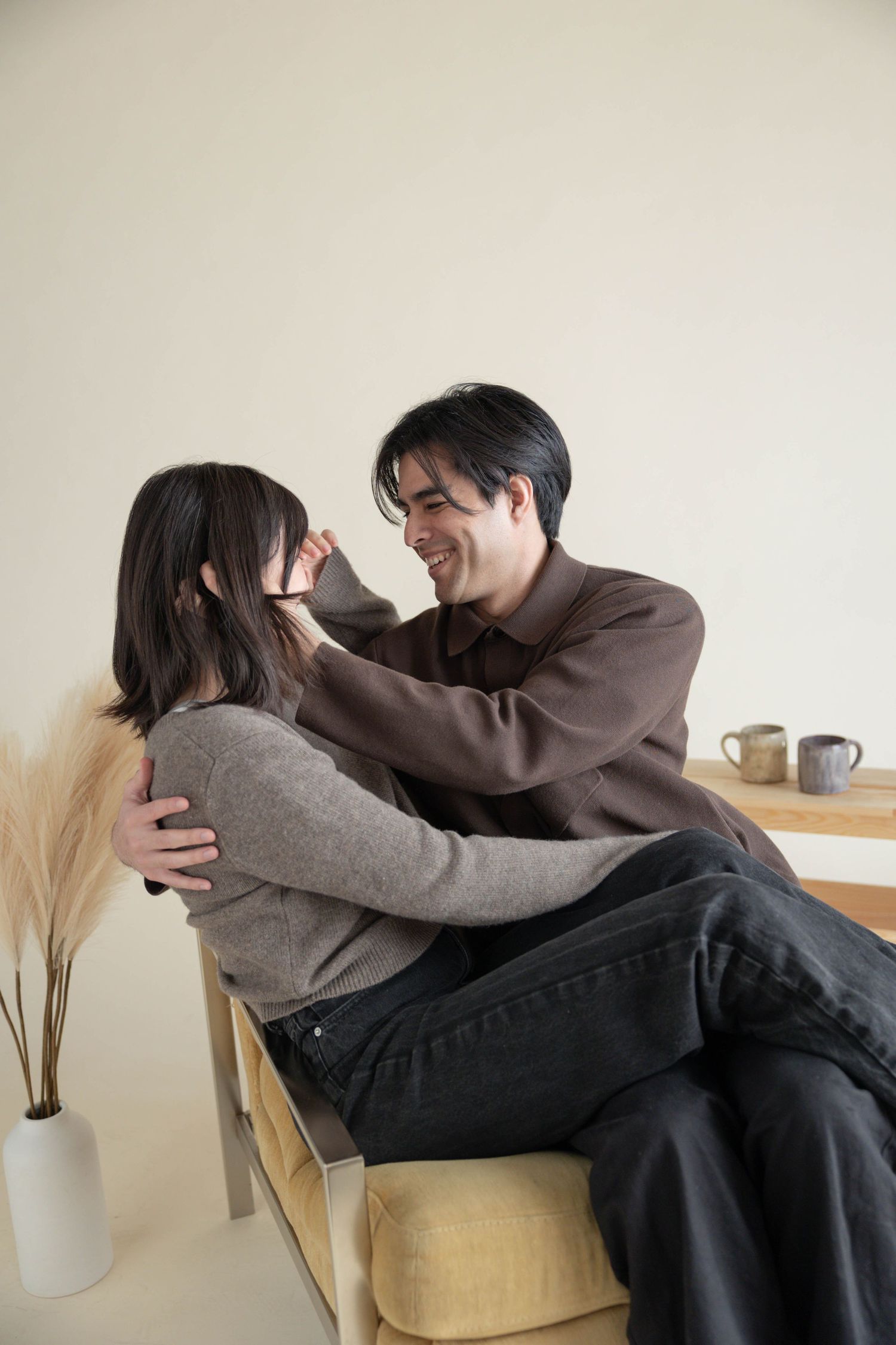 A smiling man gently brushes hair from a woman's face while they sit together on a couch in a cozy room. Couples Portraits San Diego Ca